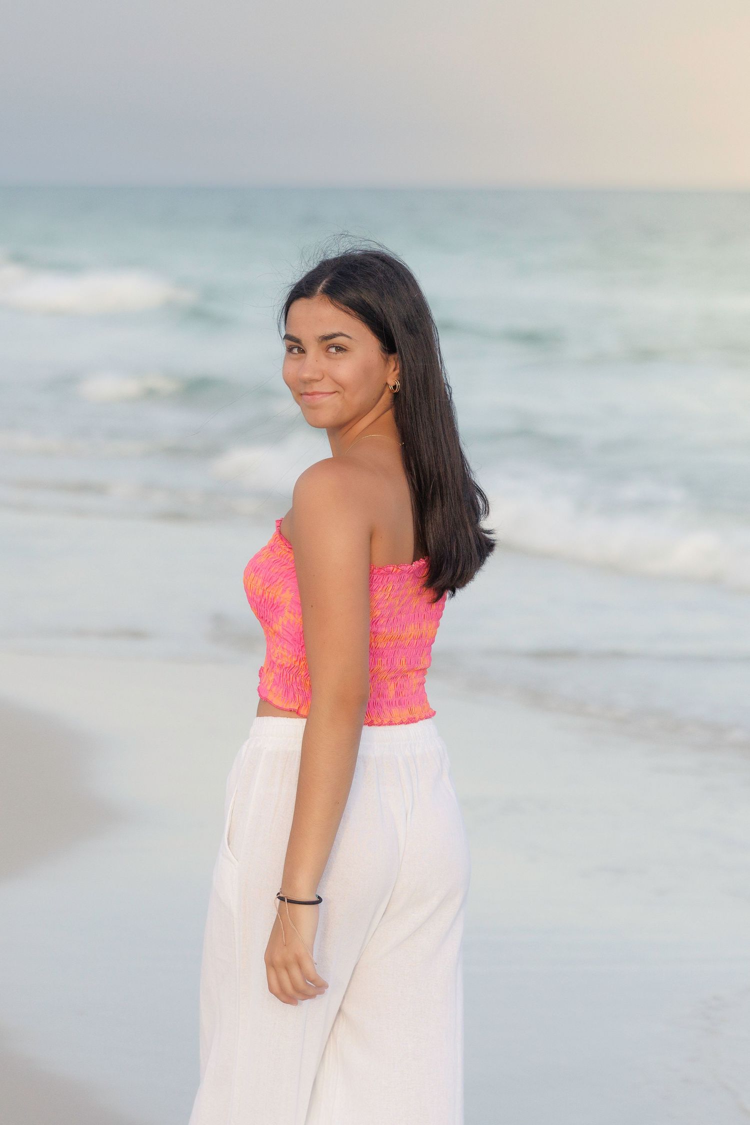 Beachside portrait session with flowing white dress and neon coral top against ocean waves and sand.