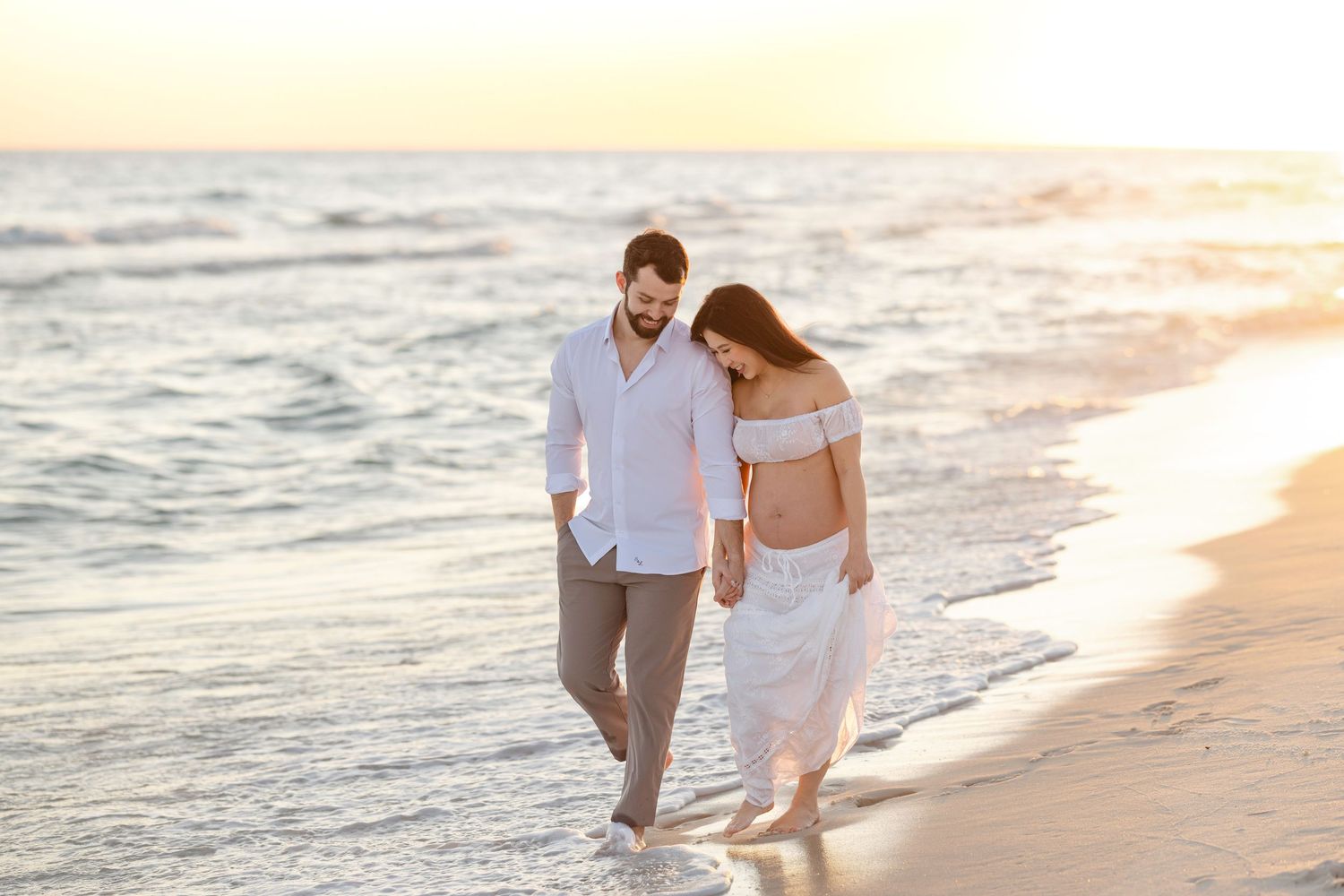 A couple walks along a sandy beach at sunset while waves gently roll onto the shore.