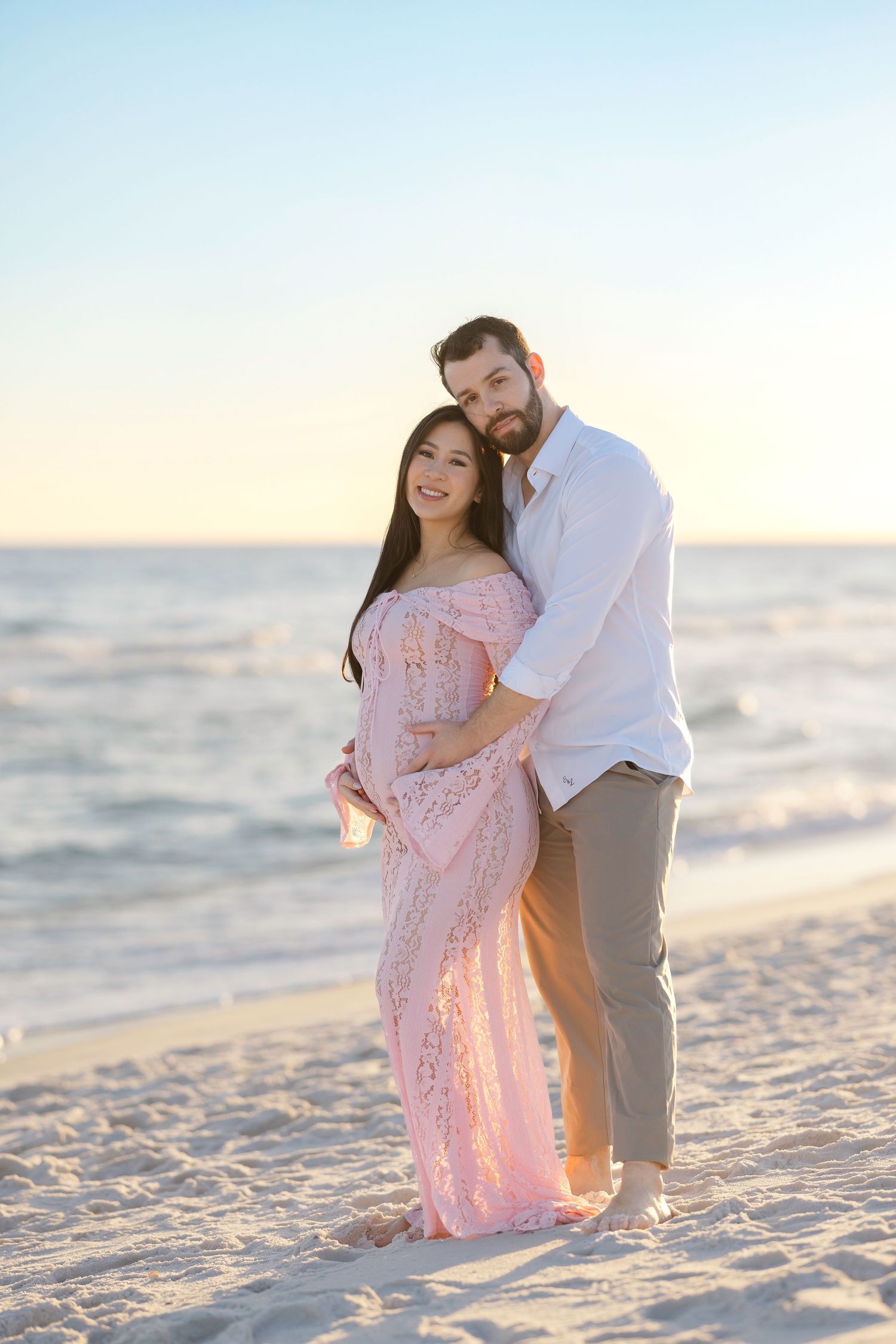 A romantic couple embraces on a sandy beach at sunset with the ocean waves in the background.