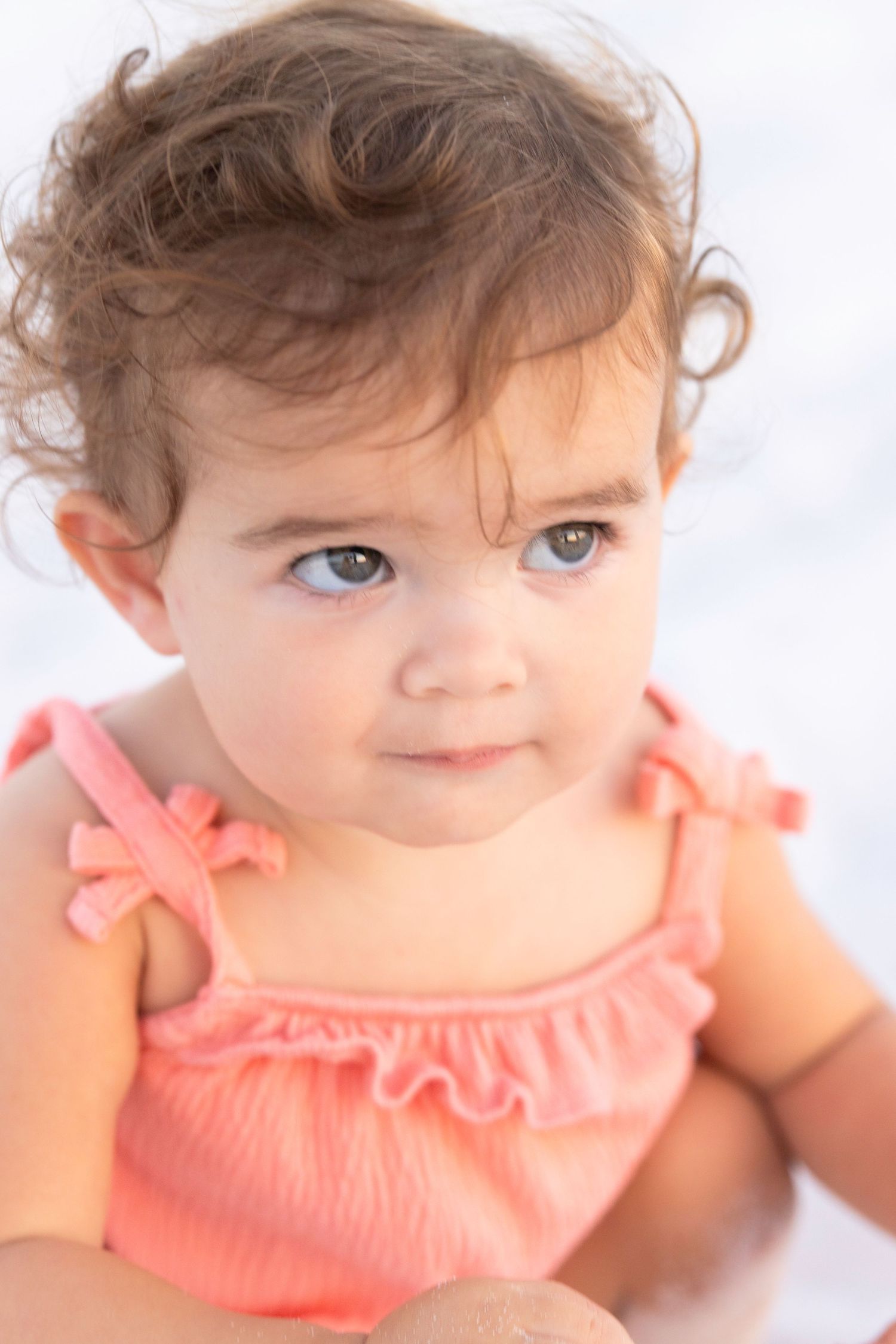 A toddler in a coral ruffled top with curly hair looks directly at the camera.