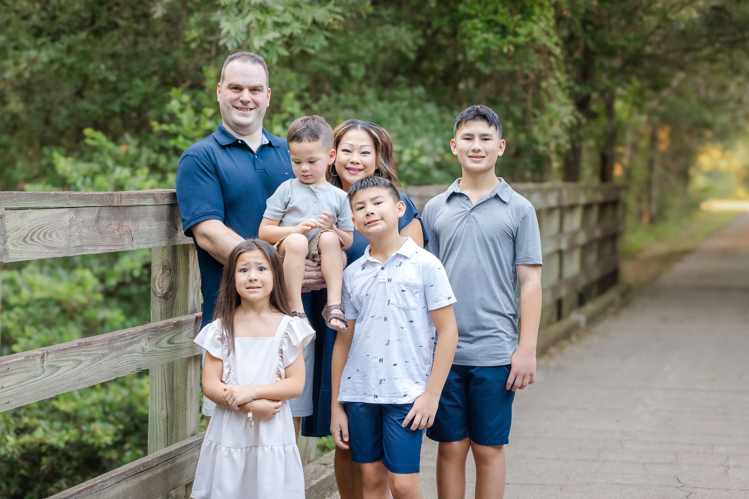 A family of six stands together on a wooden bridge surrounded by lush greenery on a sunny afternoon.