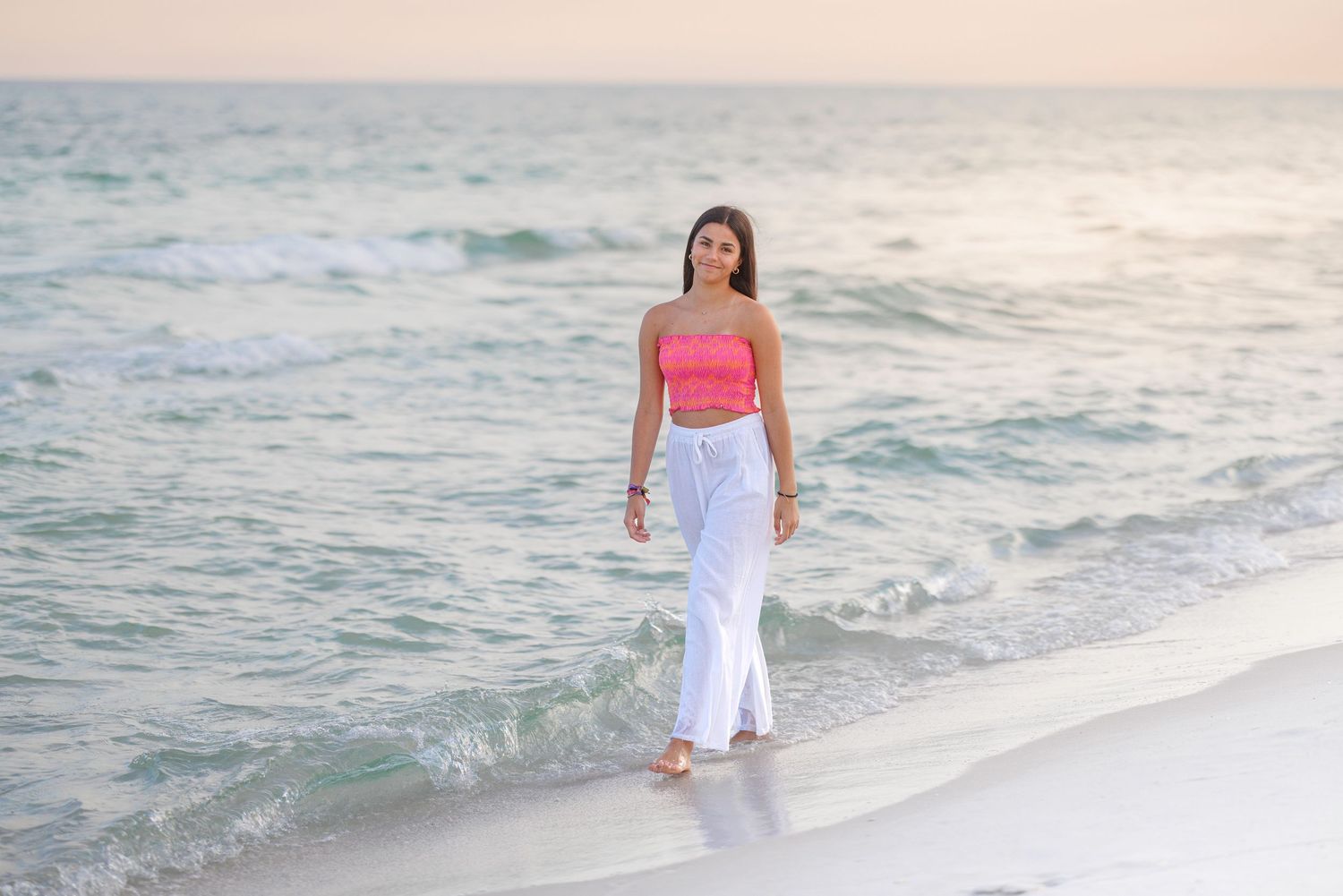 Person walking along shoreline wearing white pants and coral top during golden hour.