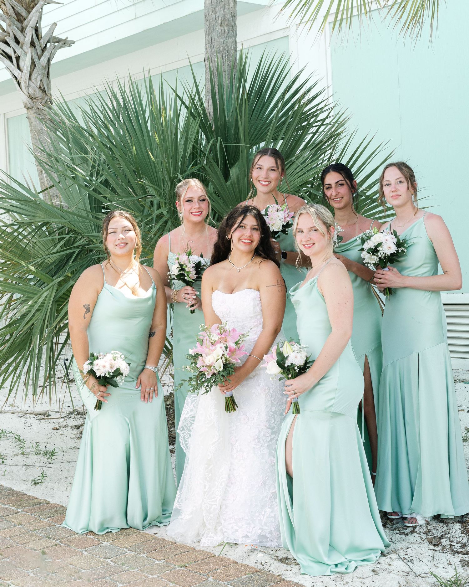 Wedding party in mint green bridesmaid dresses poses with bride in white lace gown near tropical foliage.