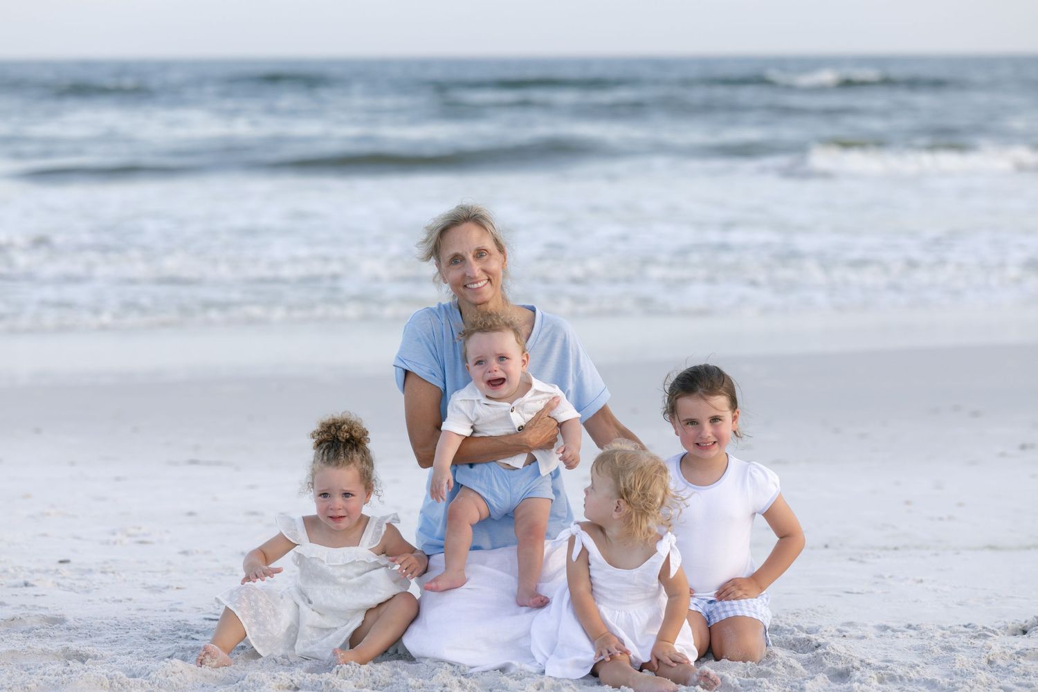 Grandmother poses with four young grandchildren on a sandy beach with ocean waves in the background.
