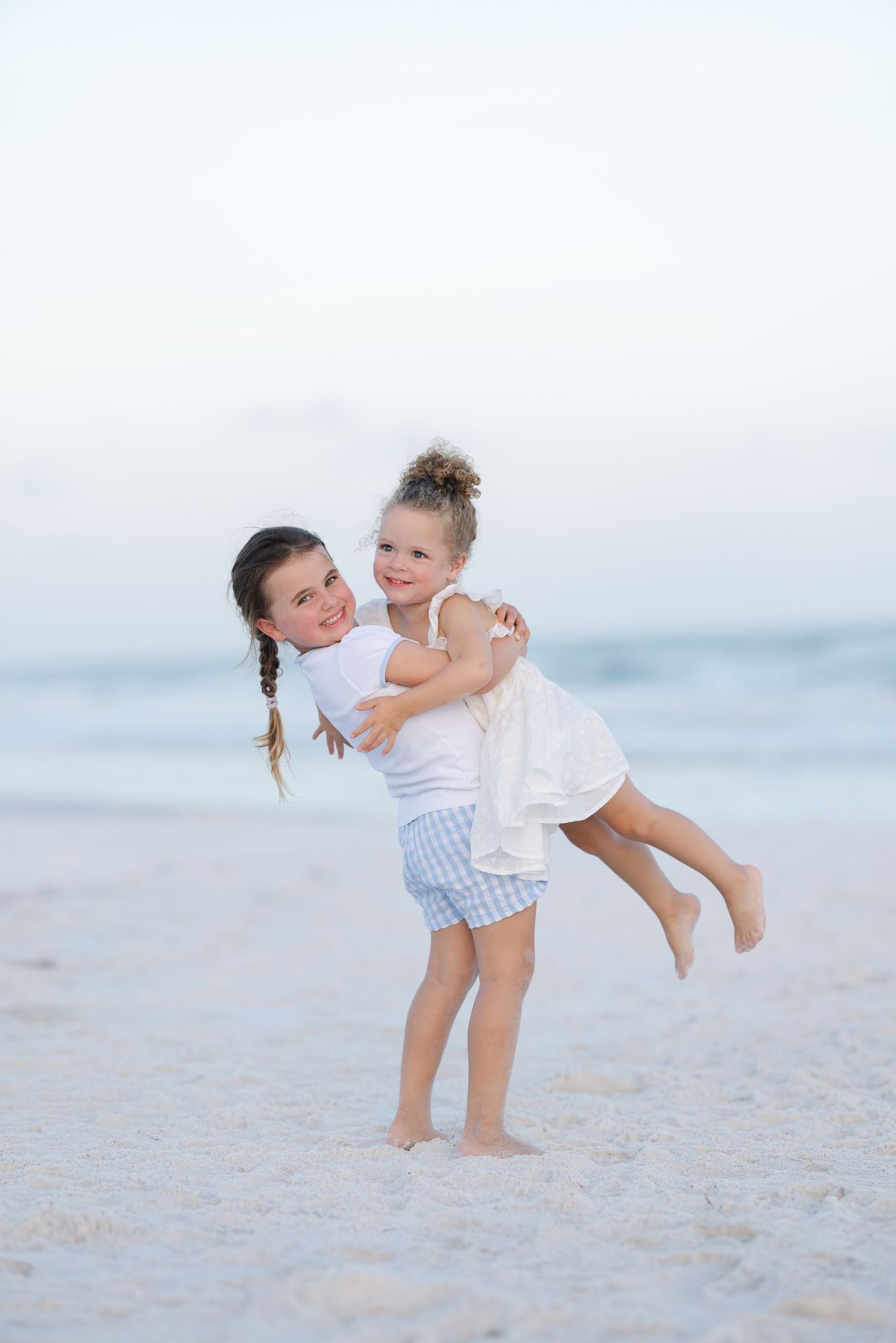 Two young girls hugging and playing on a sandy beach, one lifting the other with joy.