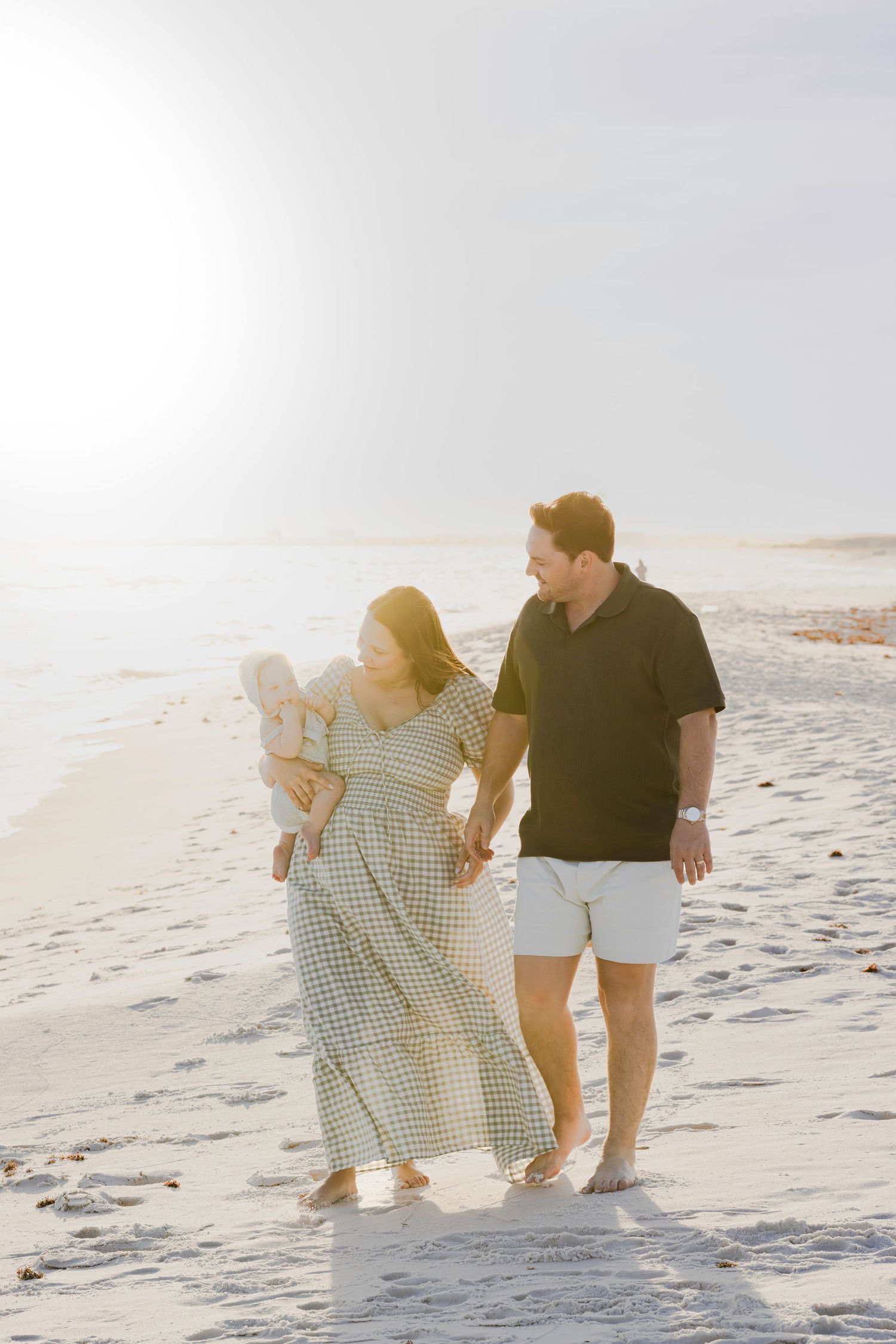 A young family walks barefoot on a sunny beach, mother holding baby in a gingham dress, father in black shirt and shorts.