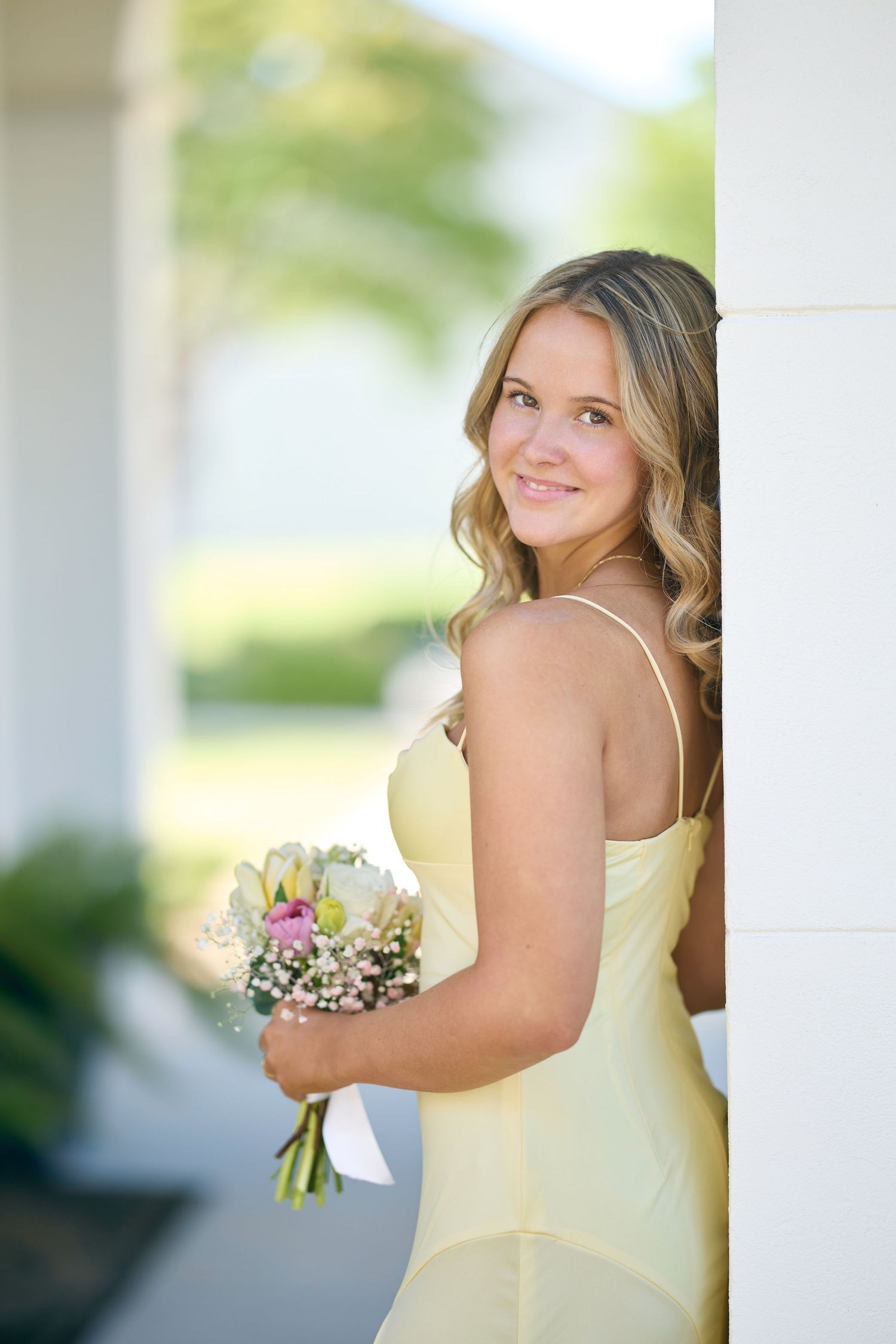 A bridesmaid in a pale yellow dress holds a white and purple floral bouquet while posing outdoors.