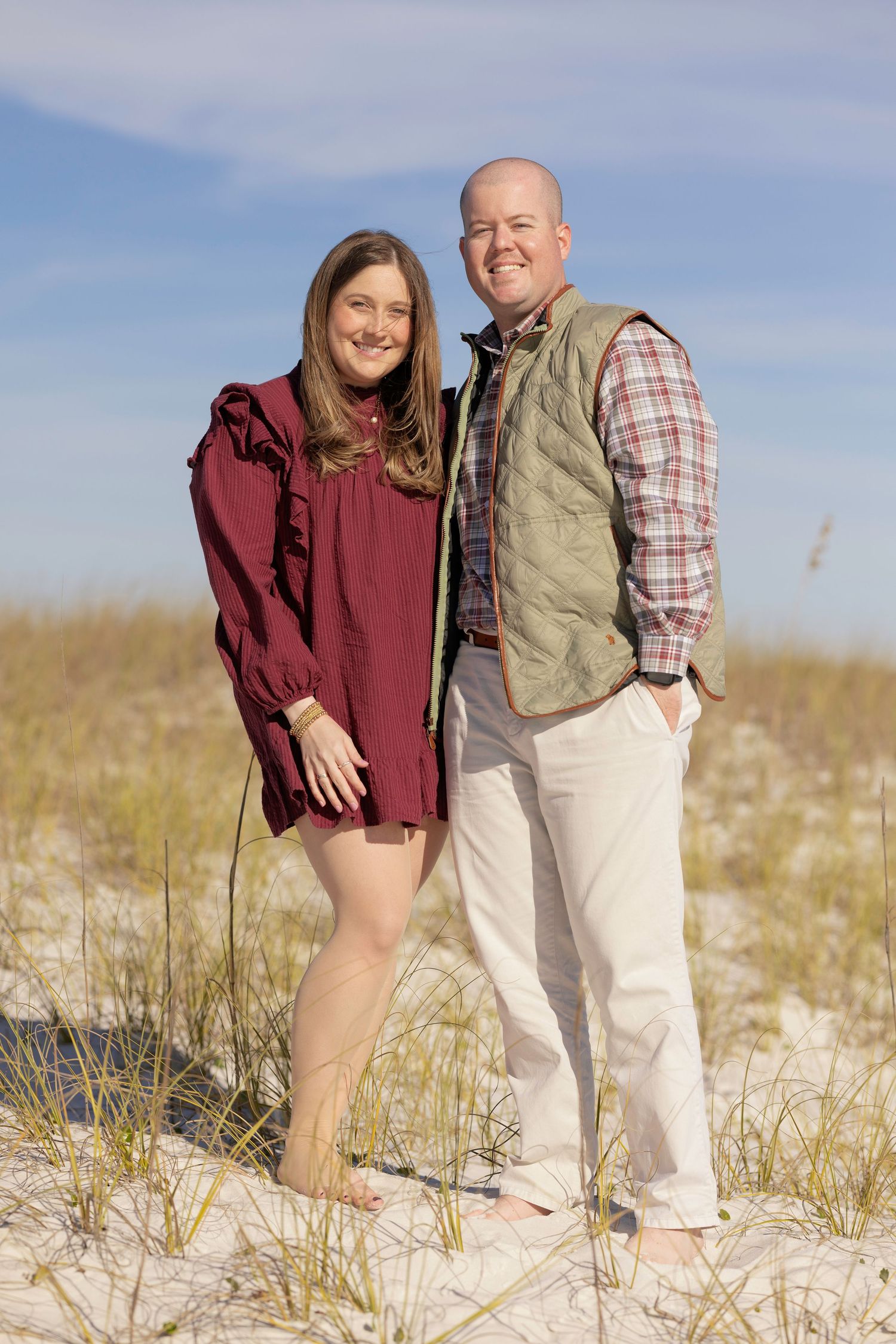 Smiling couple standing barefoot on white sand dunes, woman in burgundy dress and man in green quilted vest.