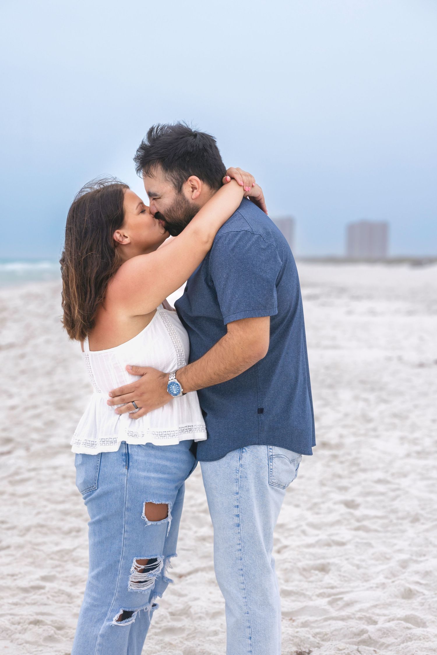 A couple shares a romantic kiss on a sandy beach, the woman in a white top and ripped jeans, the man in a blue shirt.