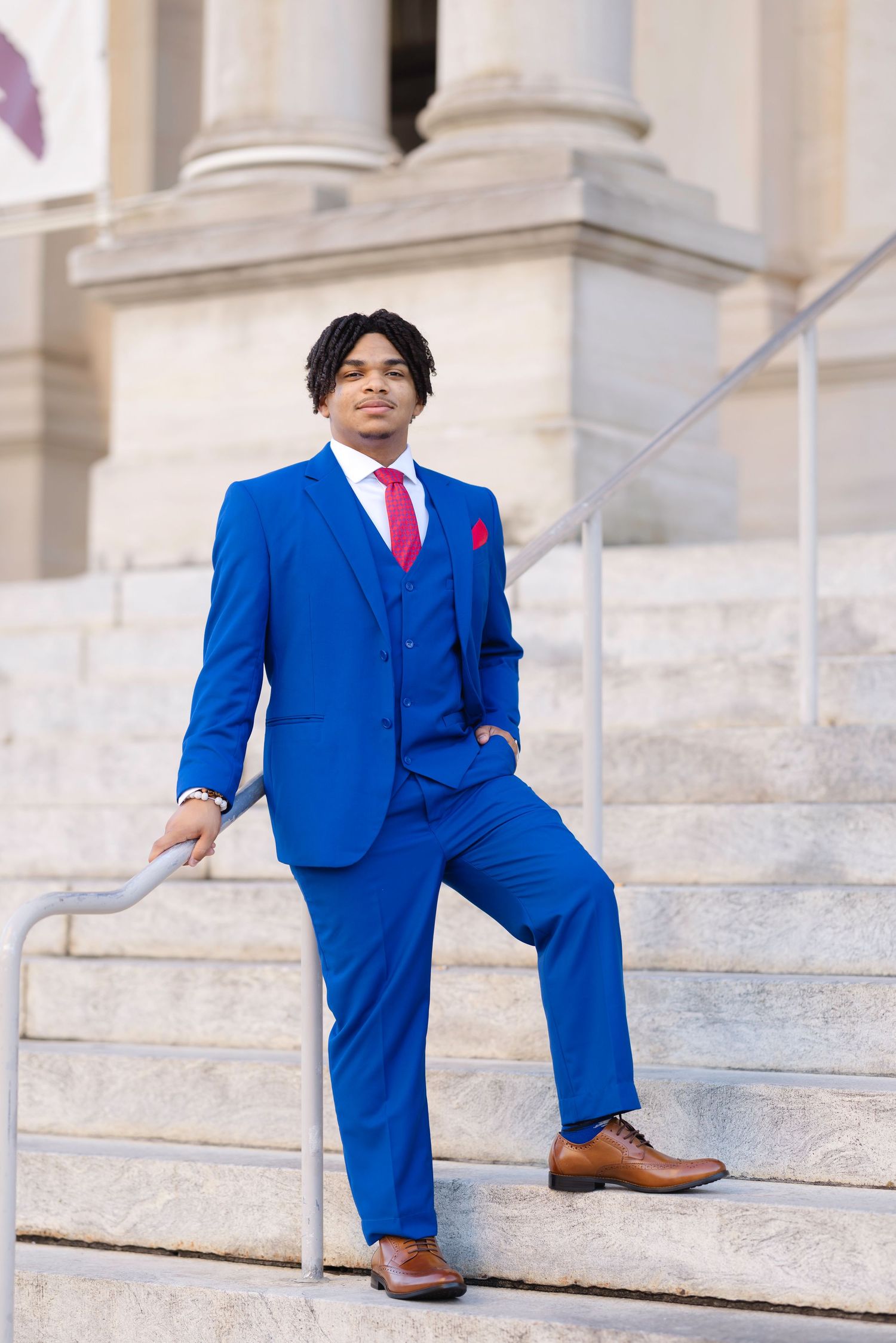 Young man in royal blue three-piece suit with red tie poses confidently on marble steps of a grand building.
