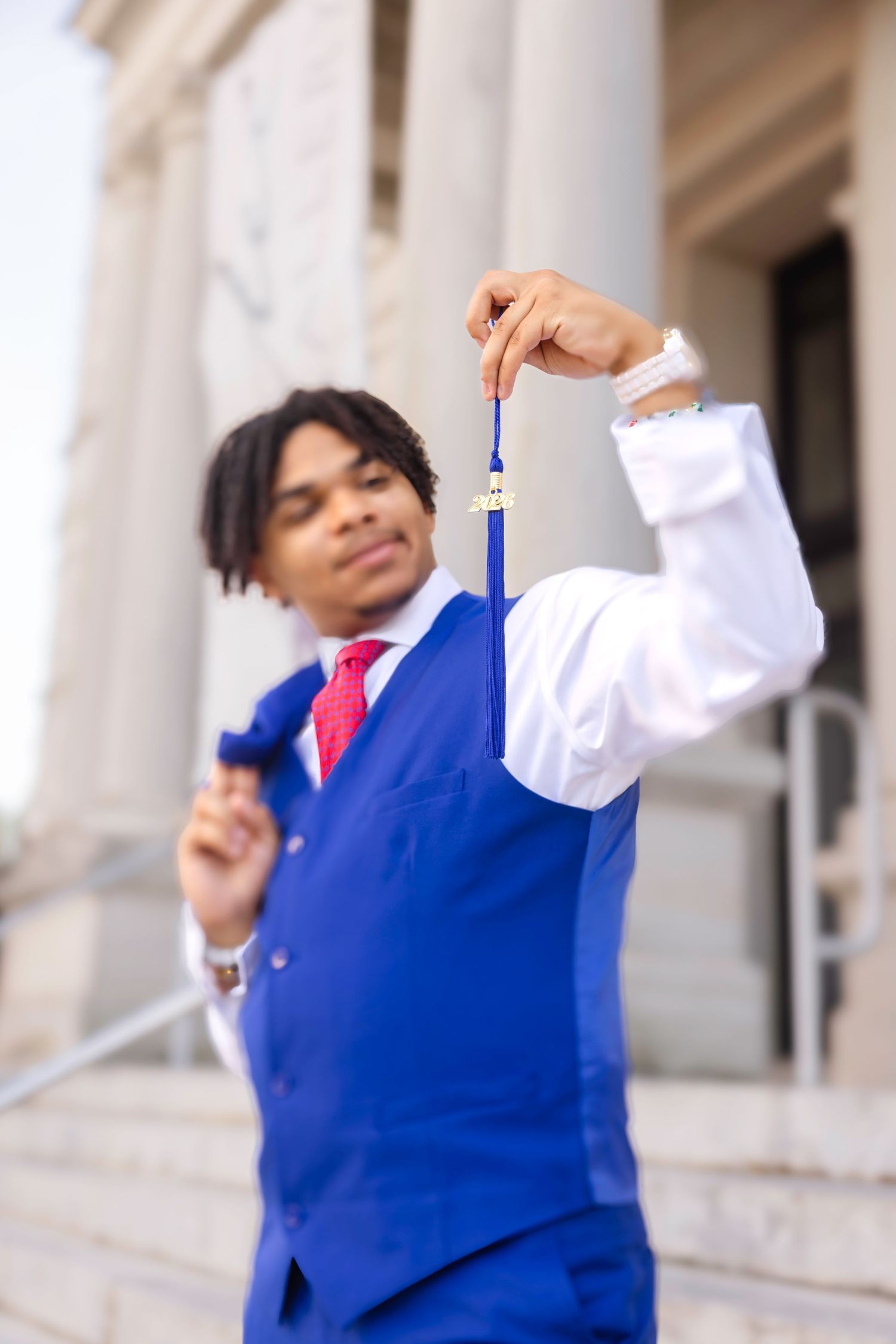 Young man in blue vest and red tie holding a graduation medal outside a formal building.