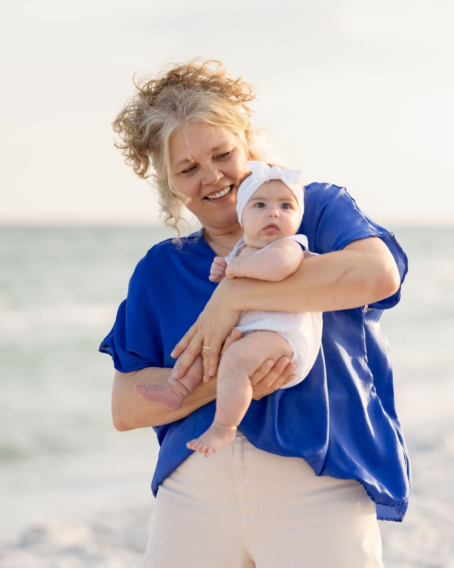 Smiling woman with curly hair holding a baby in white on a sunny beach by the ocean.