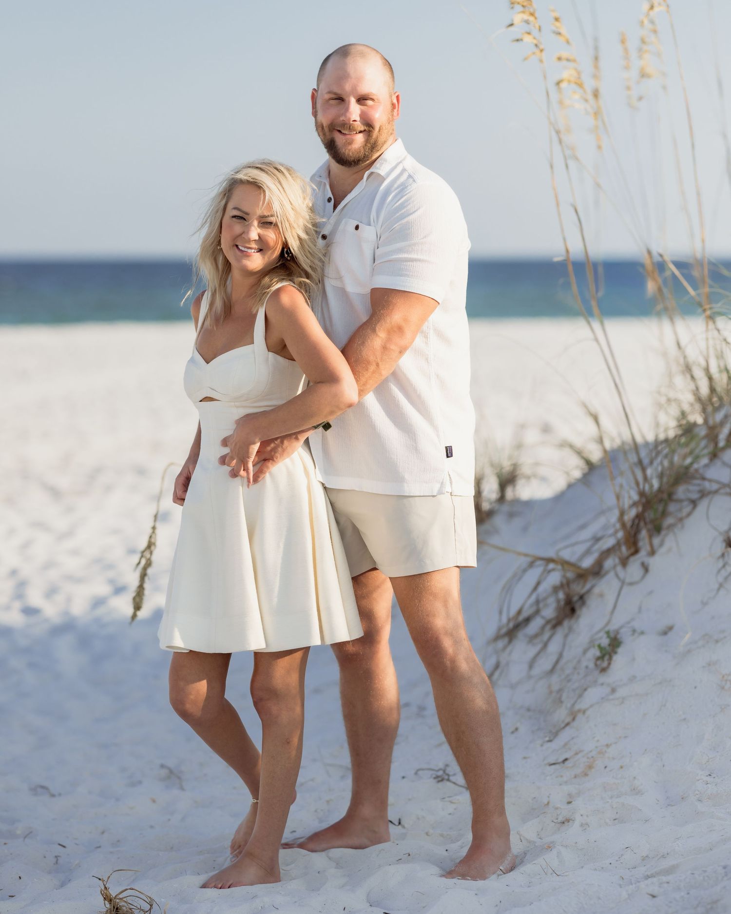 Couple in matching white outfits smiling together on a sandy beach with sea oats in the background.
