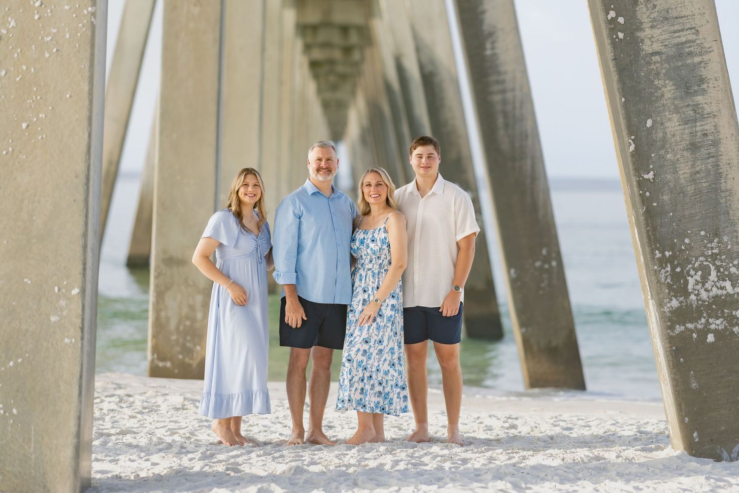 Family of four posing under a beach pier on white sand with ocean views in a sunny coastal setting.