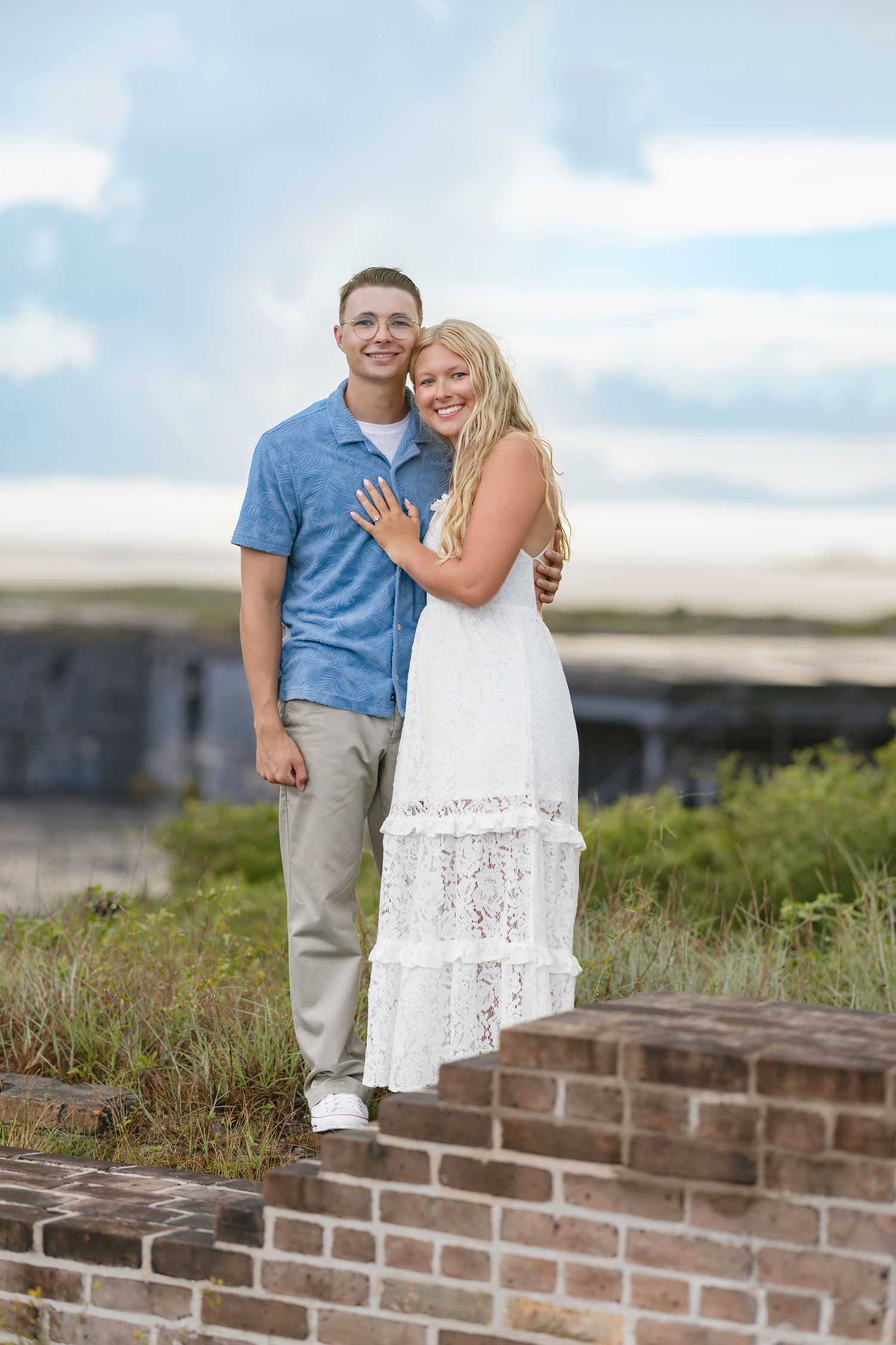 Young couple smiling and embracing outdoors near brick ruins, woman in white lace dress, man in blue shirt, cloudy sky.