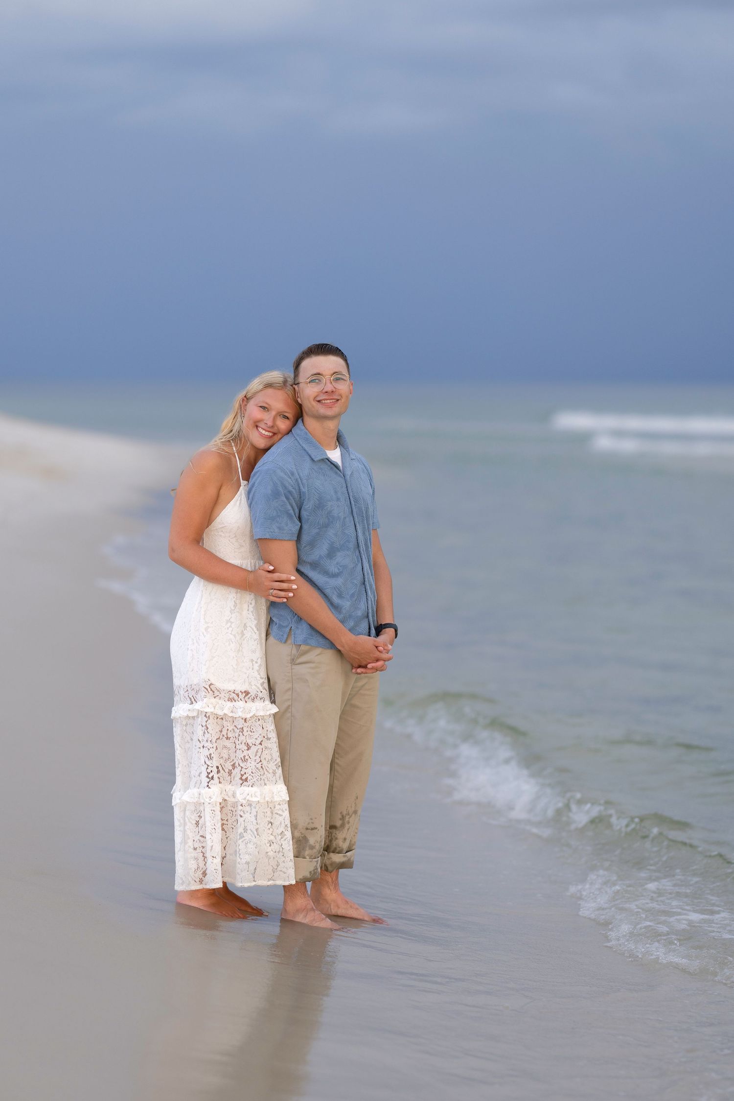 A happy couple embraces barefoot on a sandy beach, with ocean waves and cloudy sky behind them.