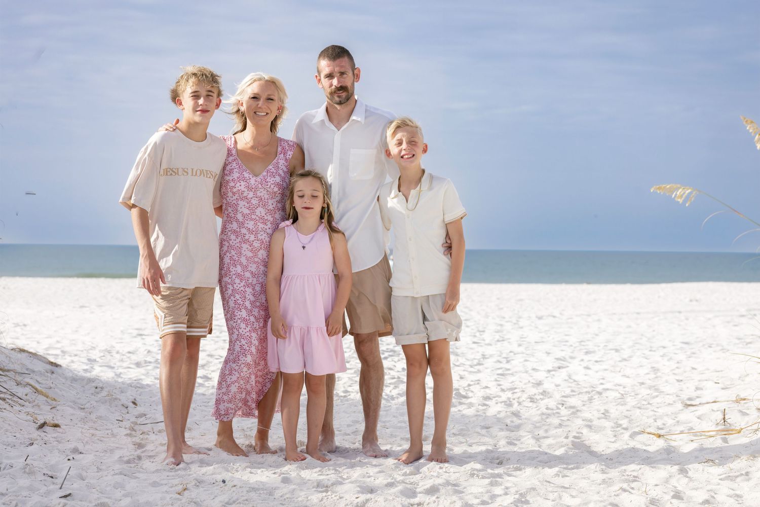 Family of five posing together on a white sandy beach with calm ocean waters in the background on a sunny day.