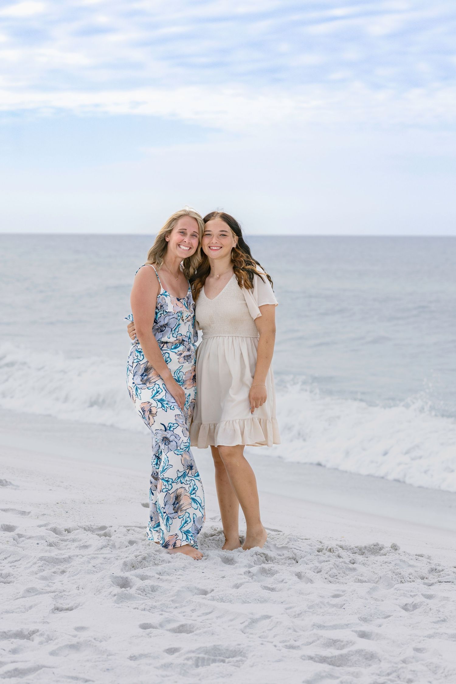 Two smiling young women in summer dresses pose together on a white sand beach with ocean waves behind them.