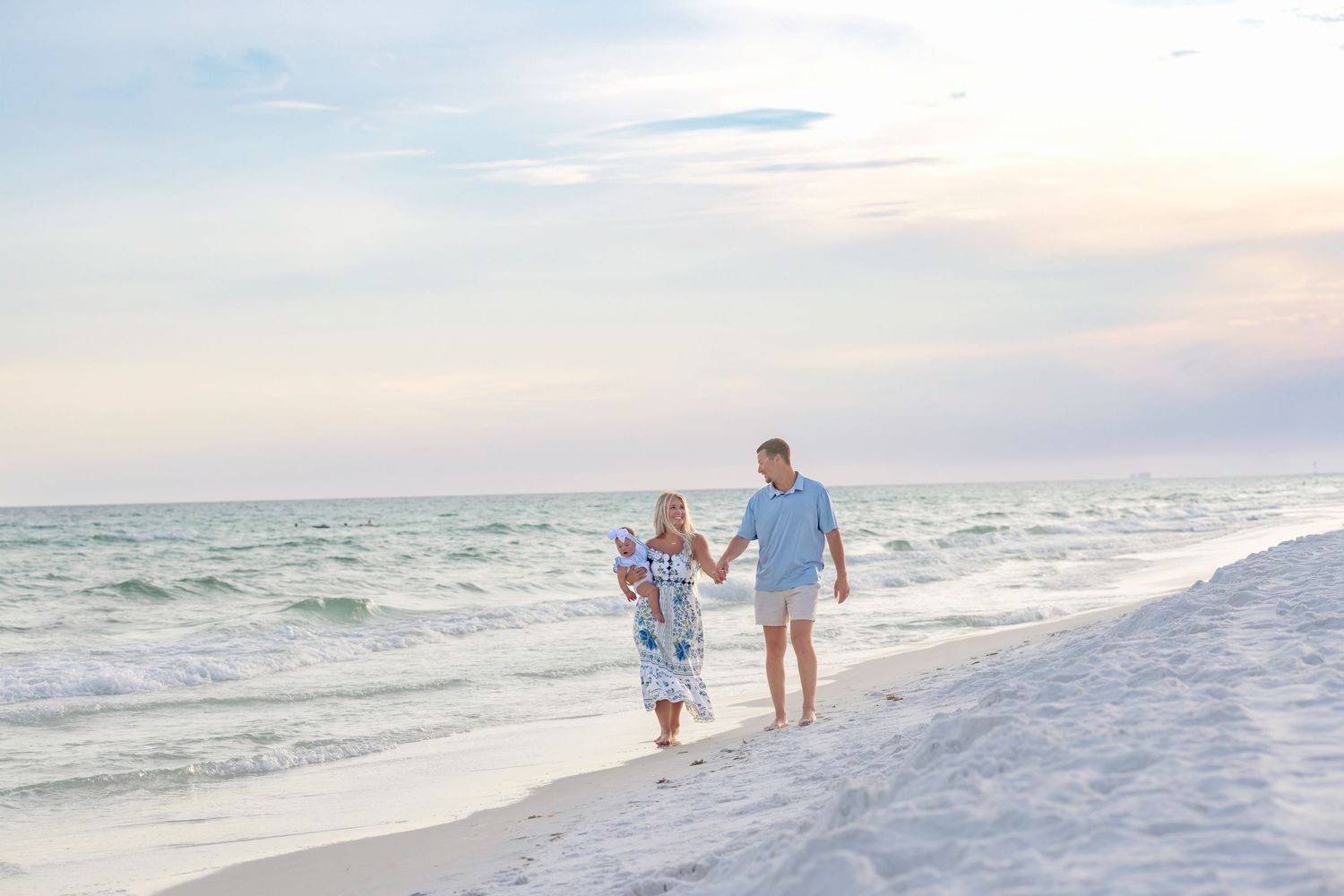 Couple holding hands walking along a white sandy beach at sunset with gentle waves and pastel sky.
