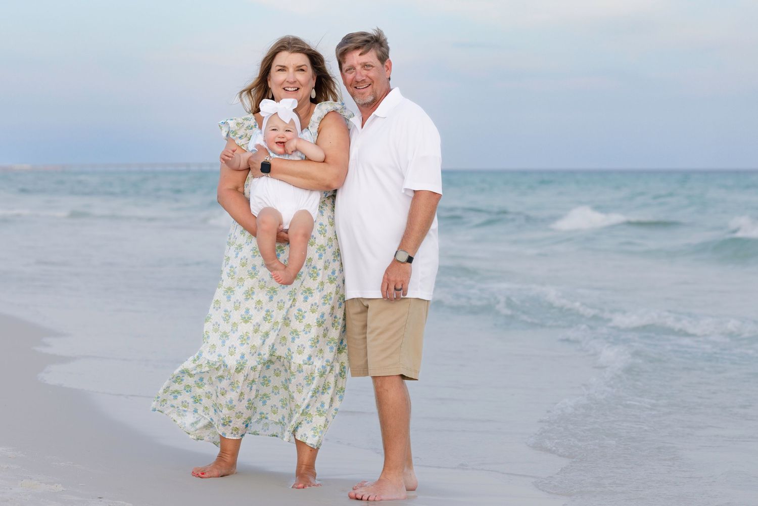 Happy family of three posing on a sandy beach, parents holding baby girl, ocean waves in background.