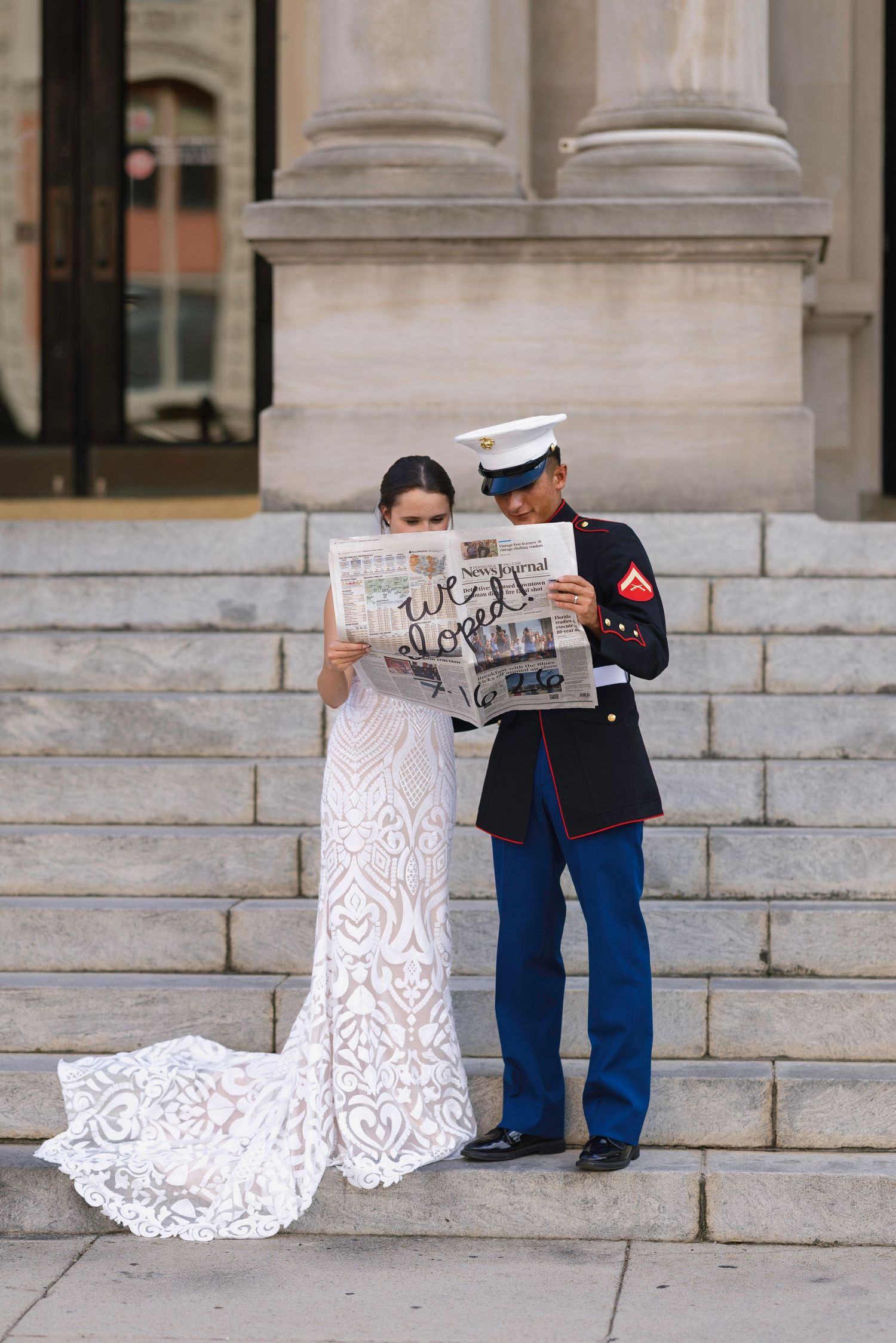 Marine groom and bride in lace wedding dress reading newspaper on stone steps of elegant building.