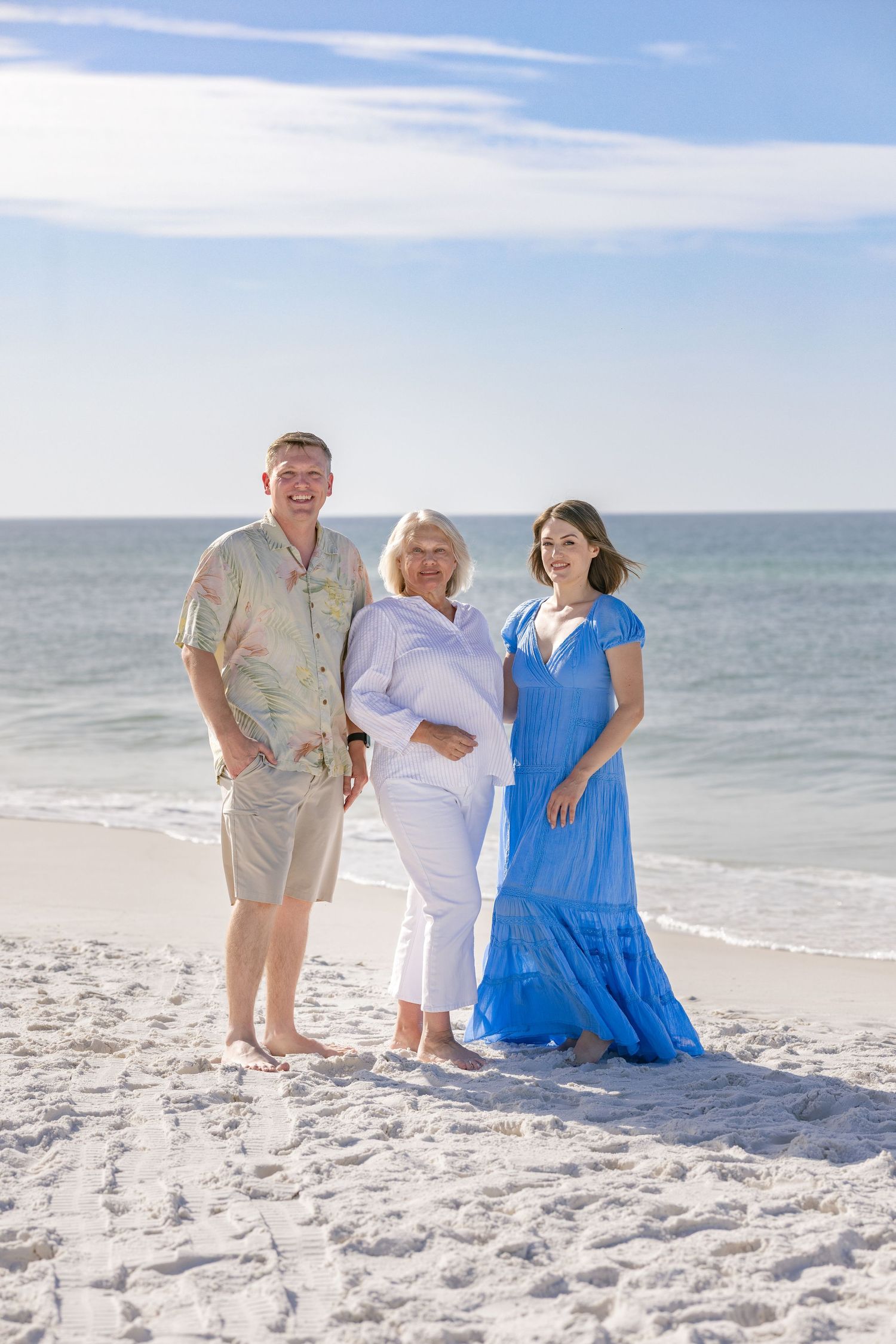 Three adults posing on sunny beach, woman in blue dress flanking older couple in casual summer attire.