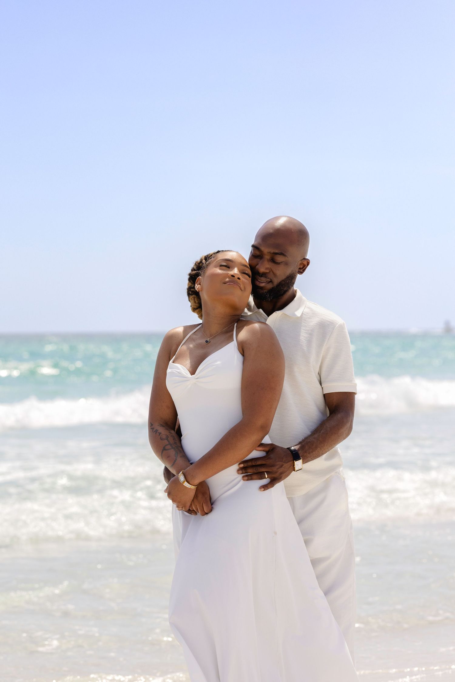 A couple in white attire embraces romantically on a sunny beach with turquoise ocean waves behind them.