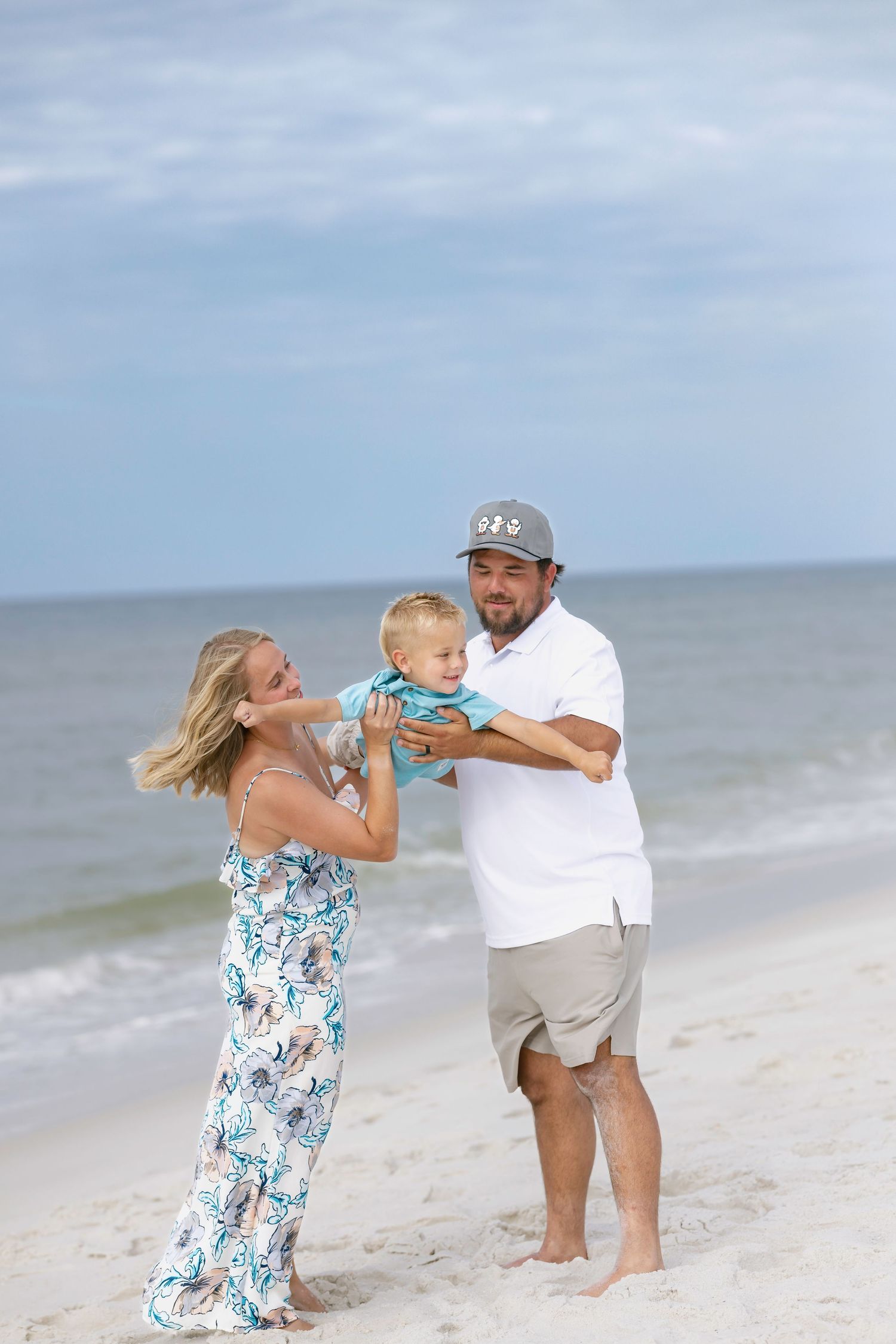 Family of three enjoying a sunny beach day, parents holding toddler on white sandy shore with ocean waves behind them.
