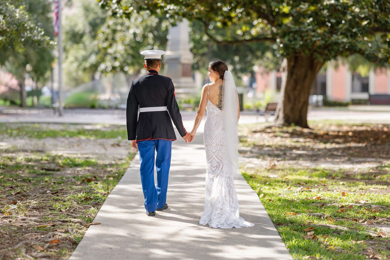 A military groom in dress uniform holds hands with his bride in a white gown as they walk along a sunlit garden path.