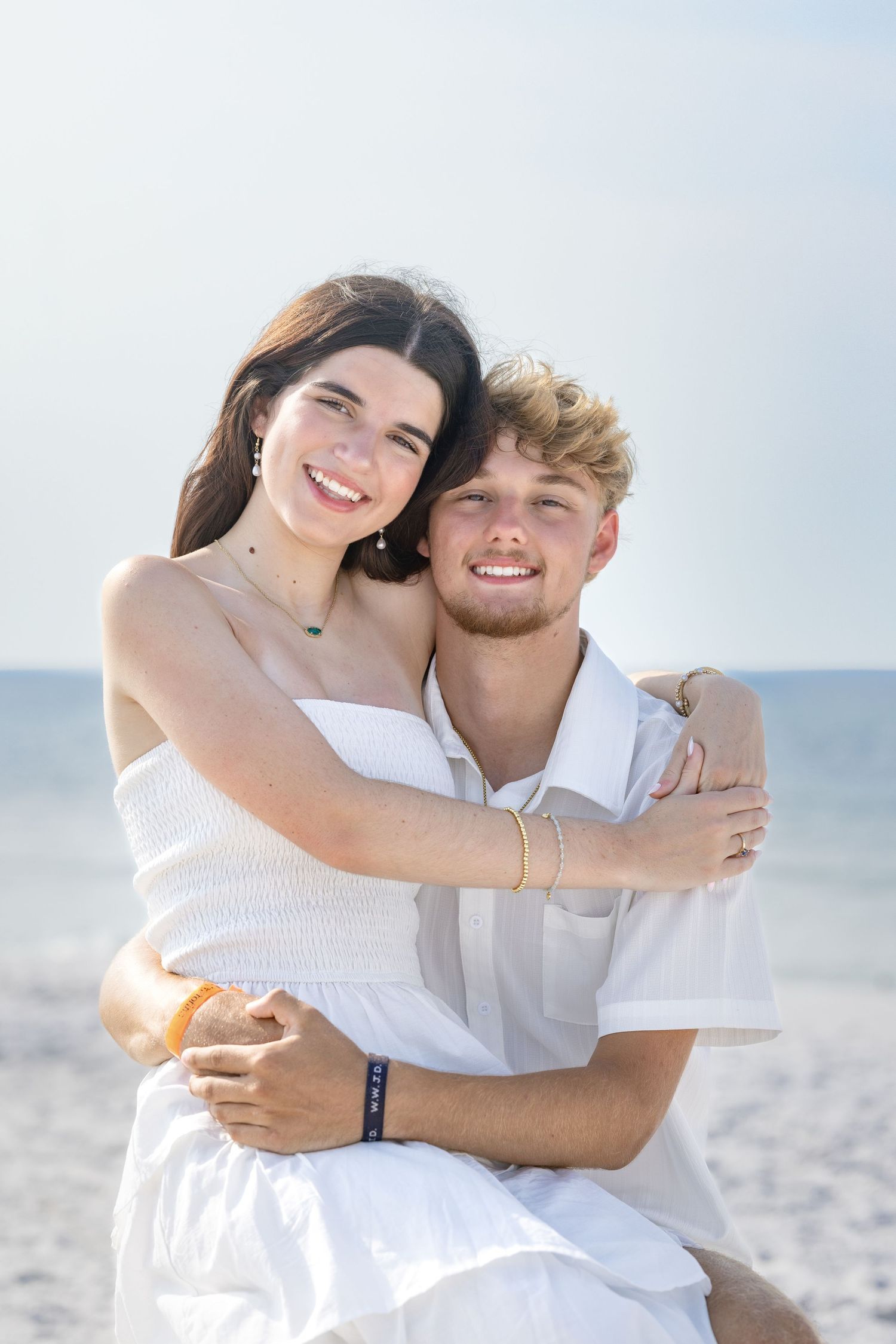 Happy young couple in white outfits embracing on a sunny beach, smiling together with ocean in background.