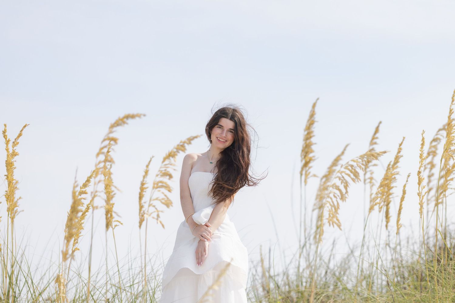Bride in white gown standing among tall sea oats on a beach, with flowing dark hair and a serene expression.