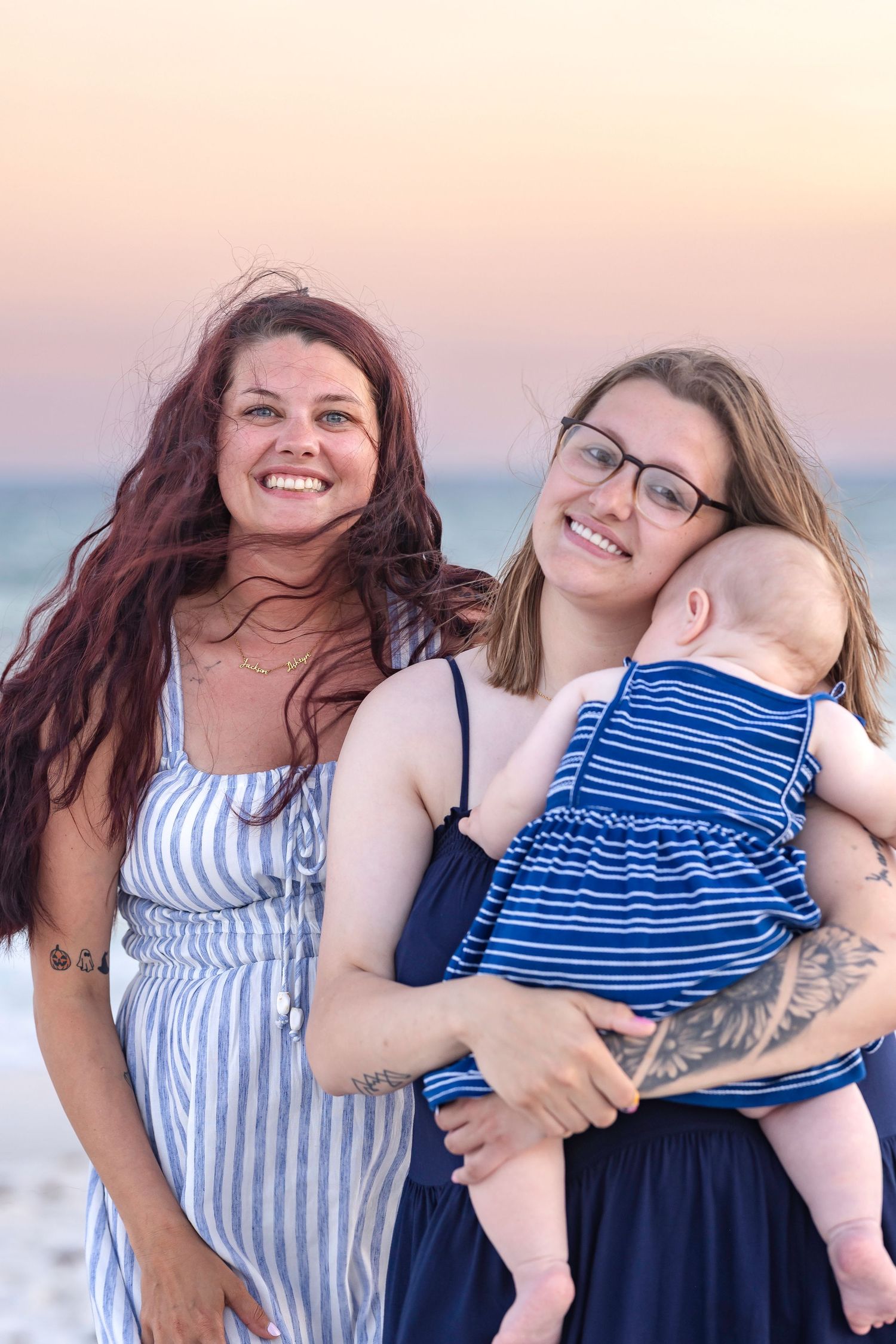 Two smiling women at beach sunset, one holding a baby dressed in a blue striped outfit.