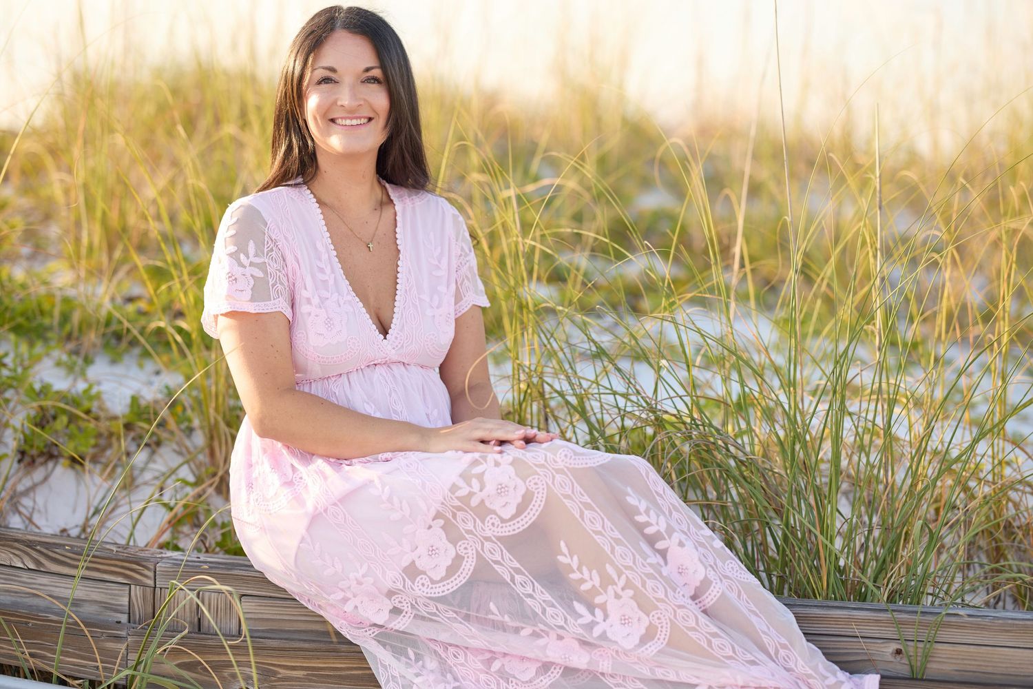 Woman in pink lace maxi dress sitting on wooden fence at sandy beach with tall grass at golden hour.