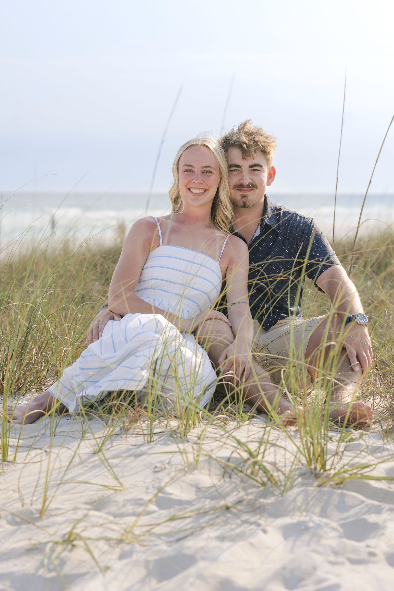 A smiling couple sits together on sandy beach dunes with sea grass, ocean waves visible in the background.