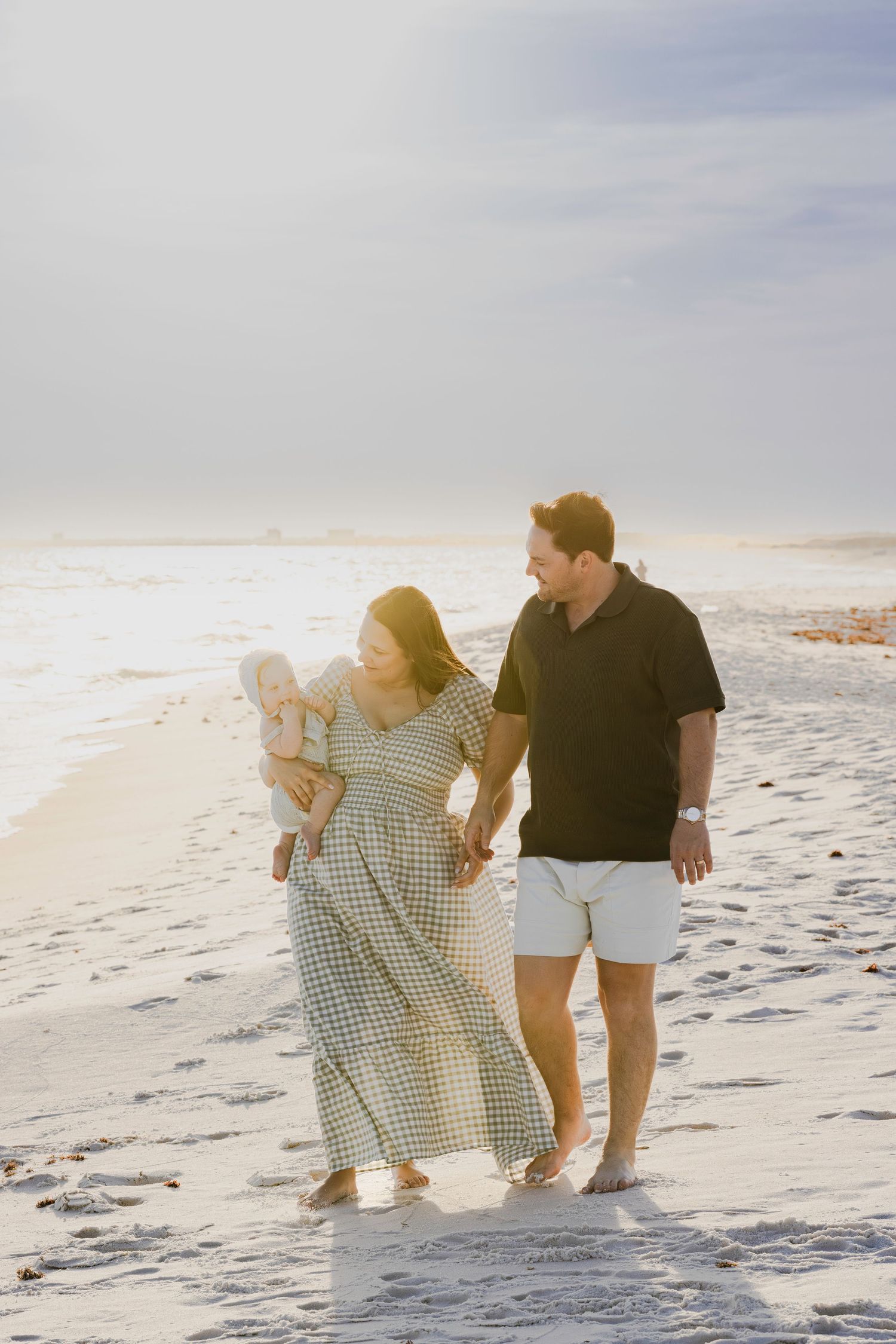 A young family walks barefoot on a sunny beach, with a woman in a plaid dress holding a baby beside a man in a black shirt.
