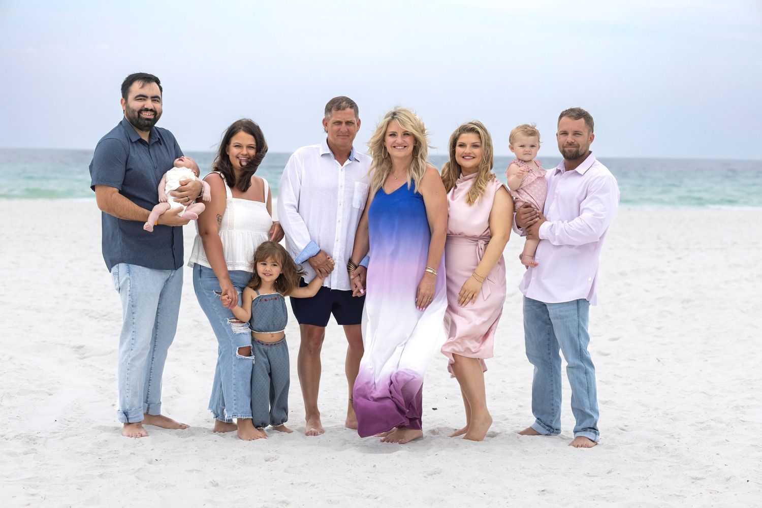 Family group portrait on sandy beach with ocean backdrop, featuring adults and young children in coordinated outfits.