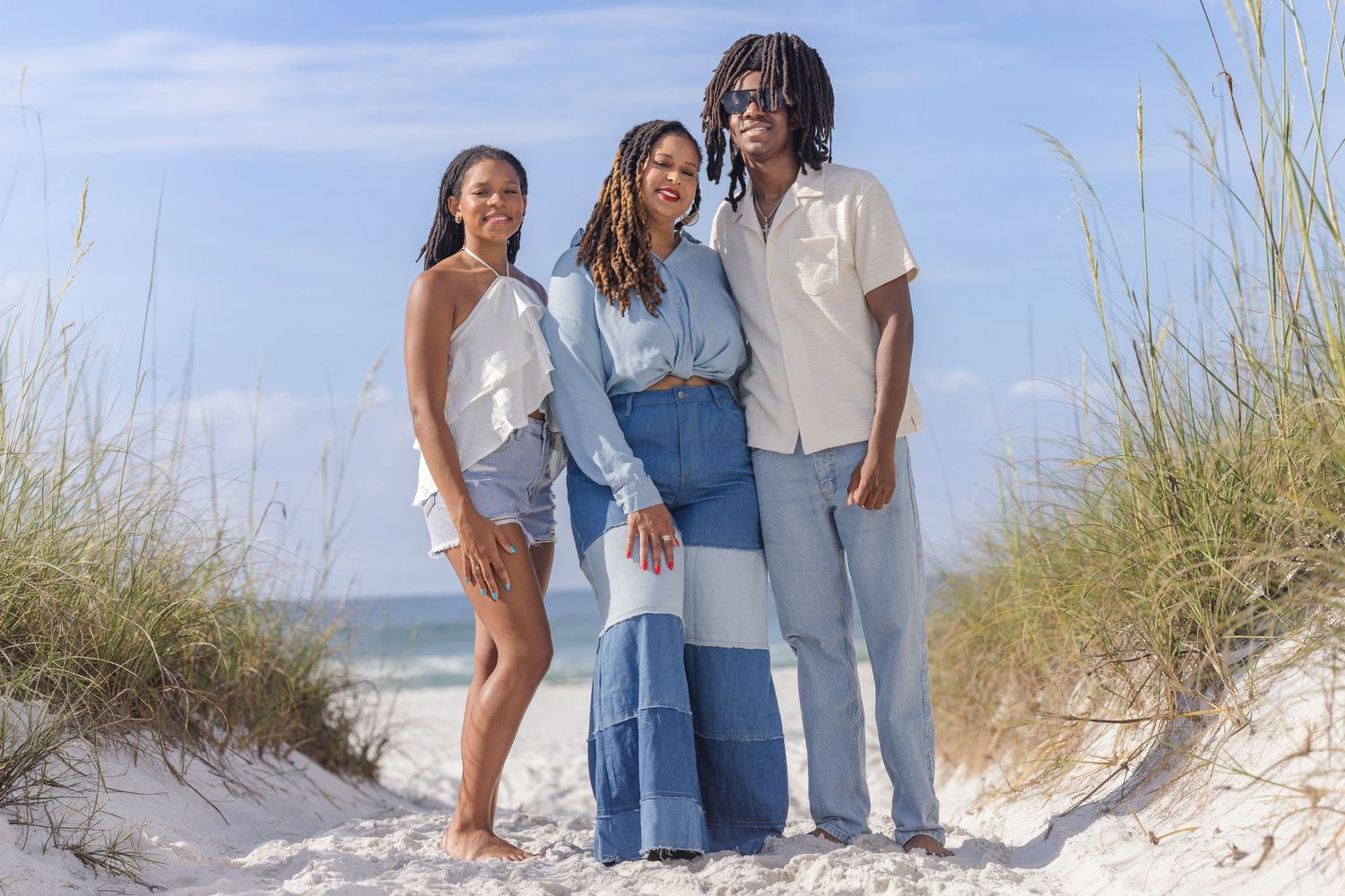 Three young adults pose together smiling on a sandy beach with ocean grass behind them.