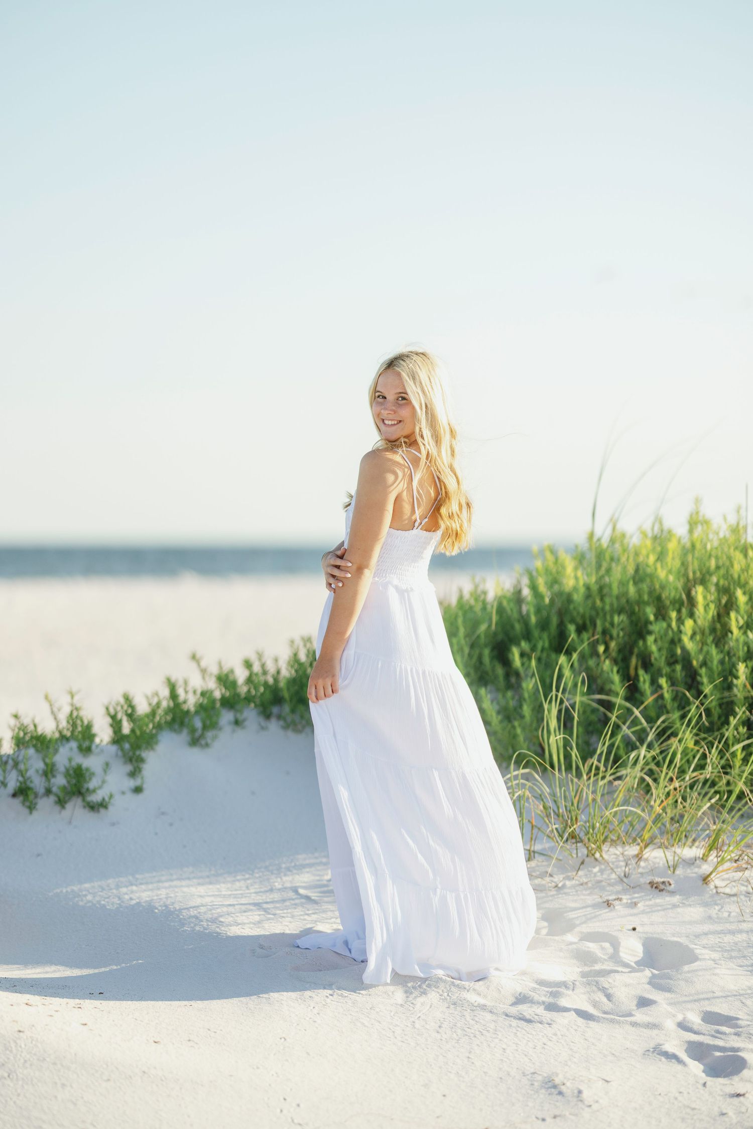 Woman in flowing white maxi dress poses gracefully on sandy beach with sea grass and ocean view at sunset.