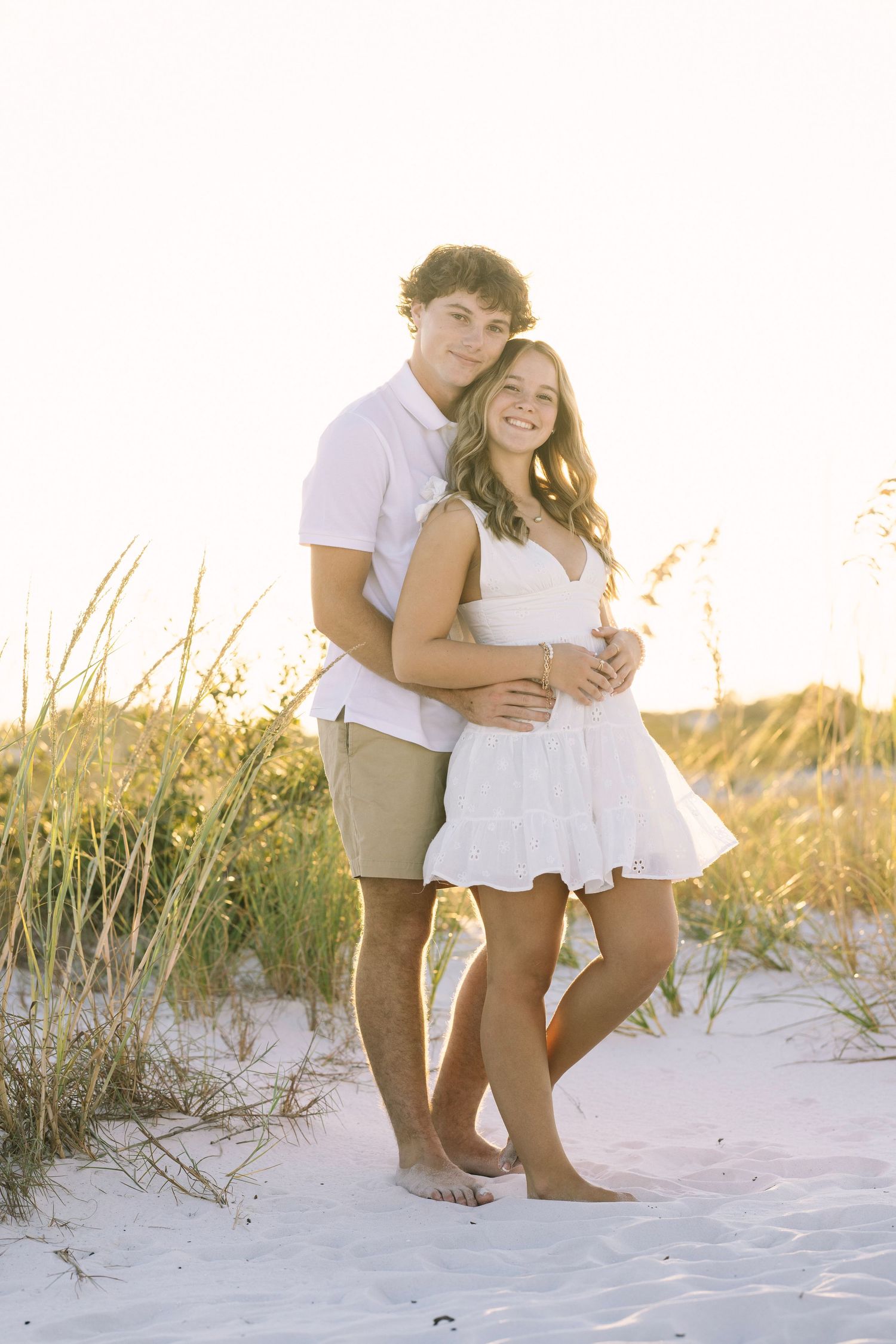 Romantic couple poses on sandy beach with sea grass during golden hour sunset.