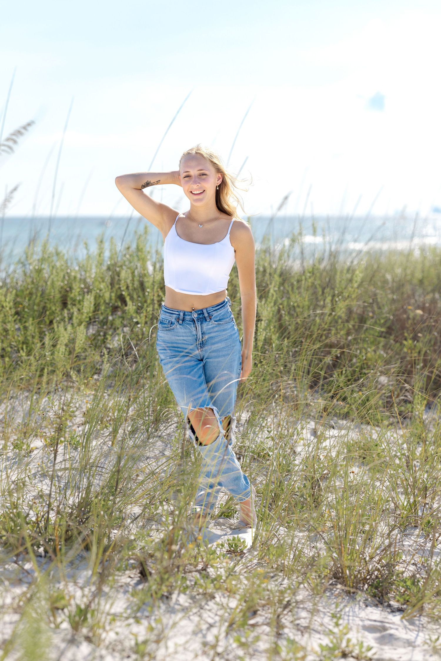Person in white crop top and ripped jeans poses casually among beach dunes with sea grass and ocean in background.