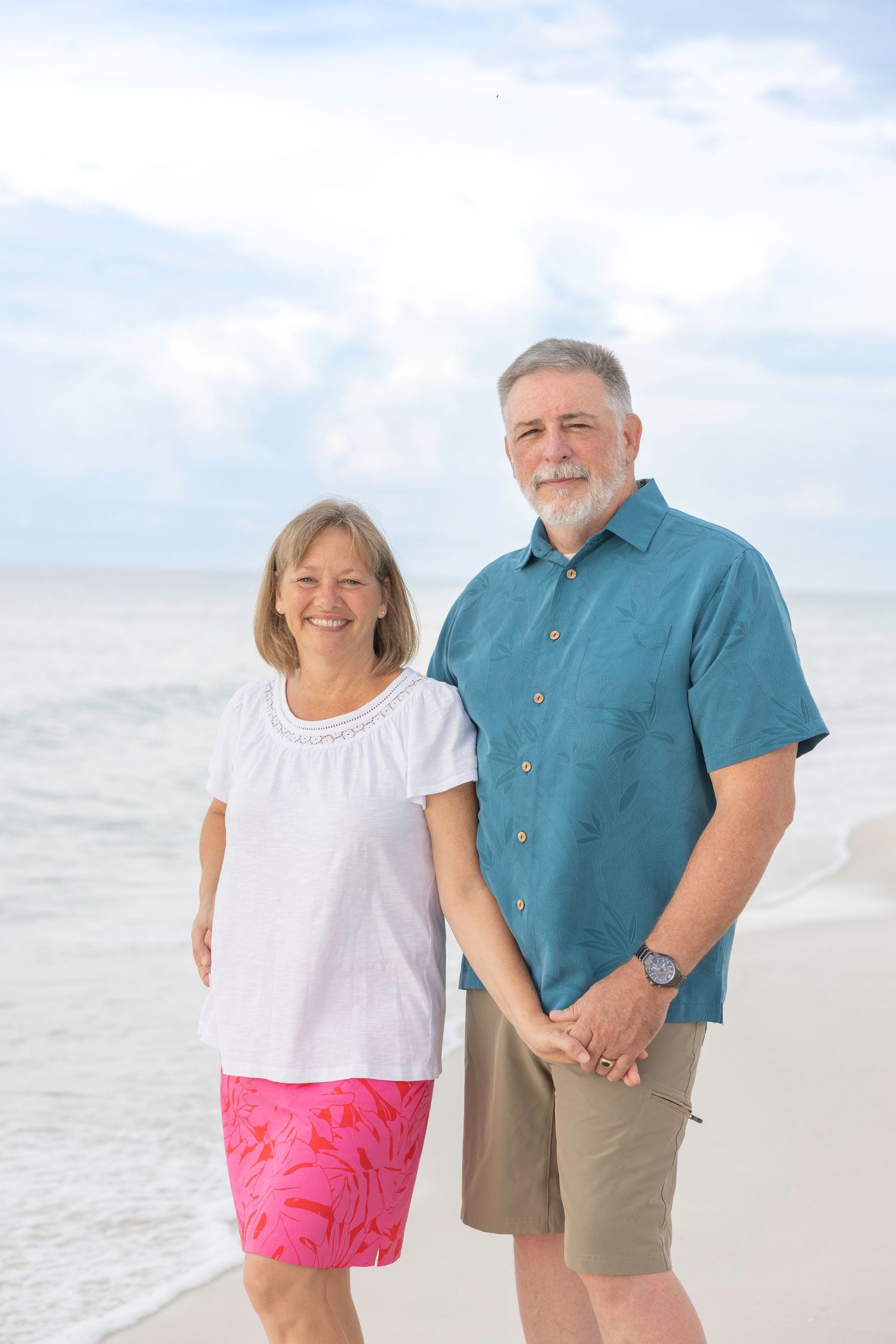 A couple poses together on a sunny beach, wearing coordinating casual outfits of teal and pink.