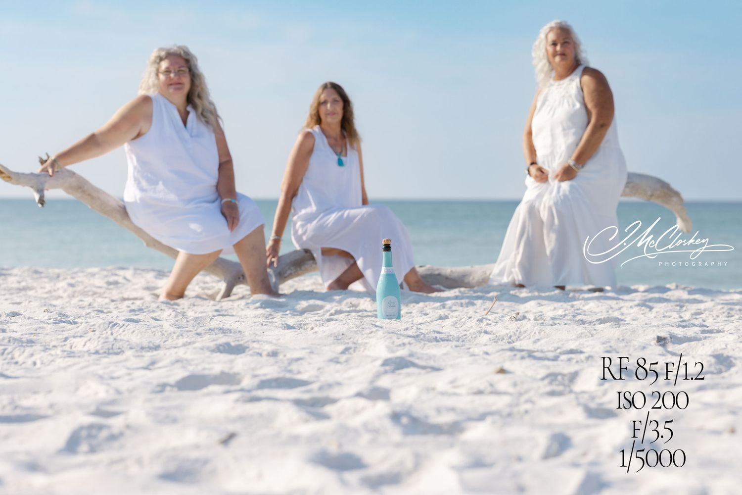 Group wearing flowing white dresses poses together on a pristine white sandy beach with ocean waves in background.