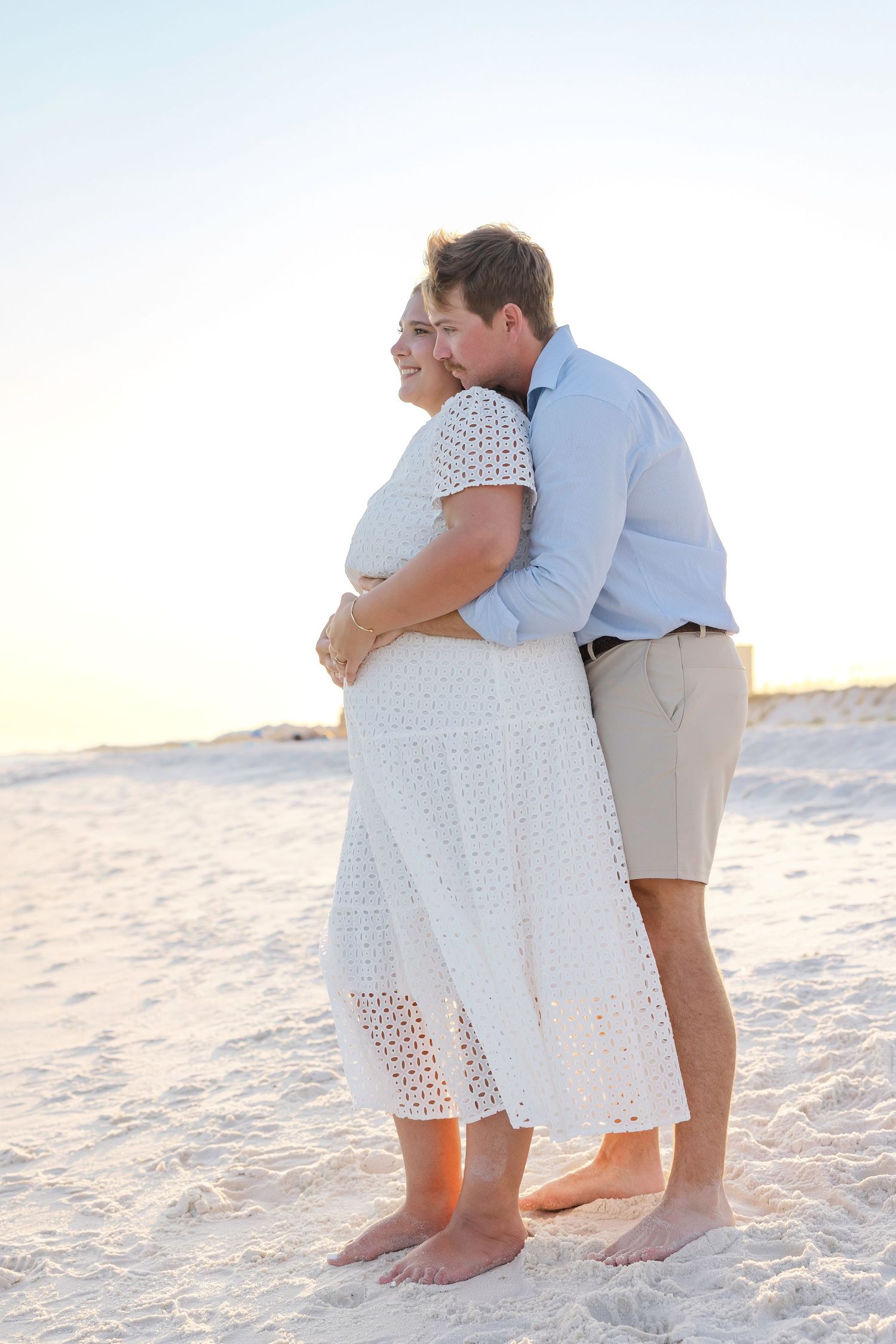 A couple shares an intimate barefoot embrace on a white sandy beach at sunset wearing light summer attire.