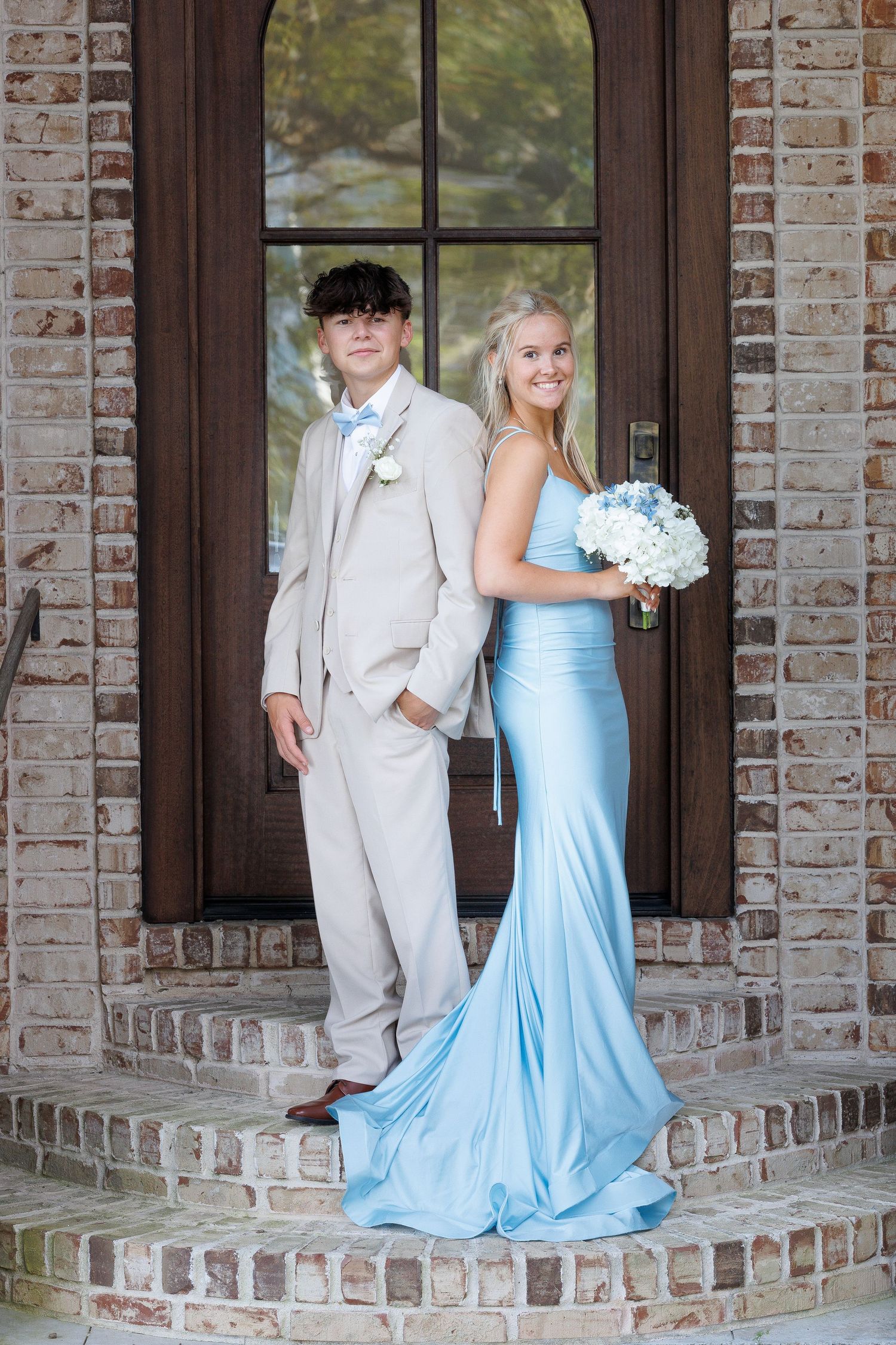 A couple poses on brick steps wearing formal attire - a light beige suit and flowing light blue gown with a bouquet.