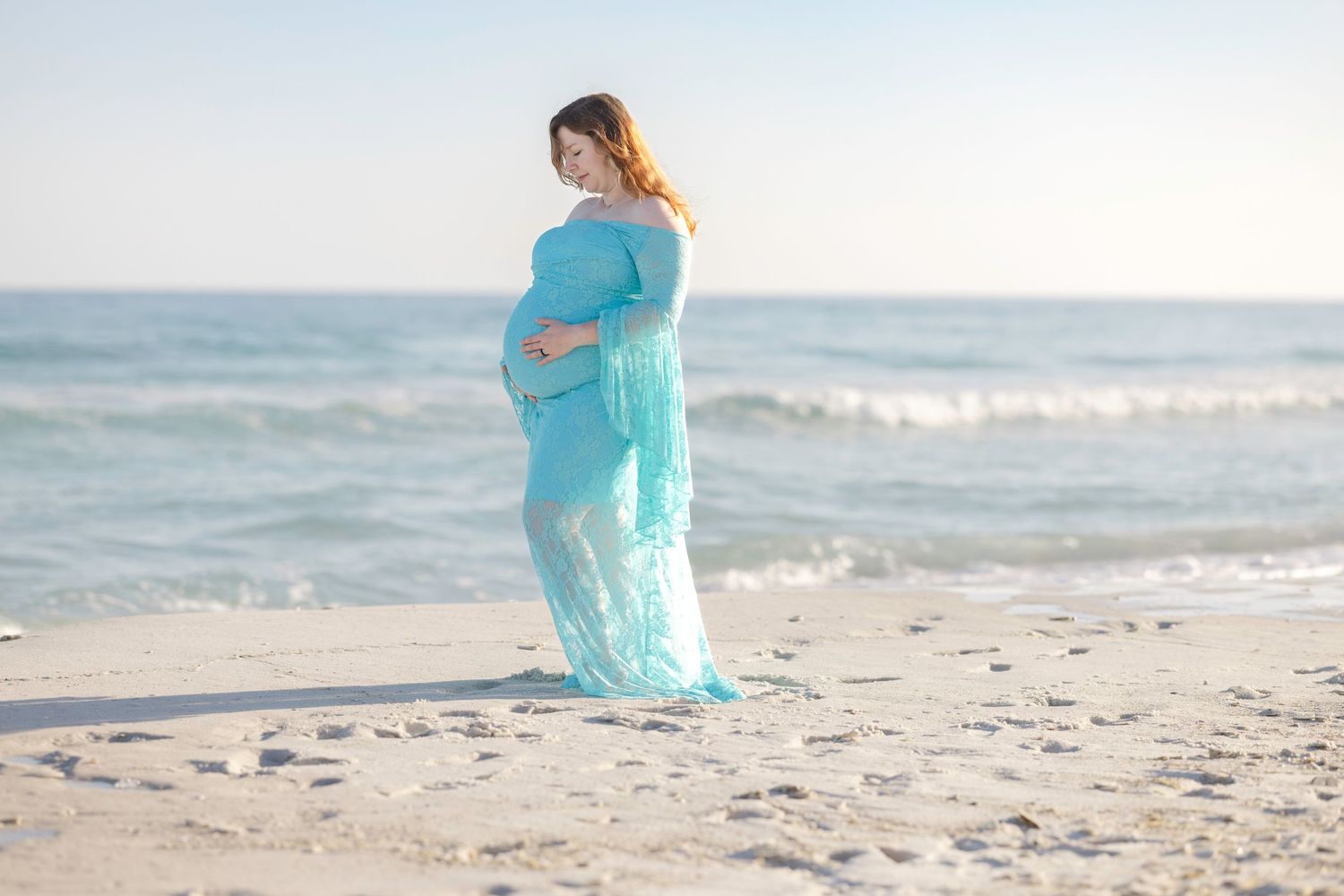 Pregnant woman in teal lace gown poses on sandy beach with ocean waves in background during golden hour.