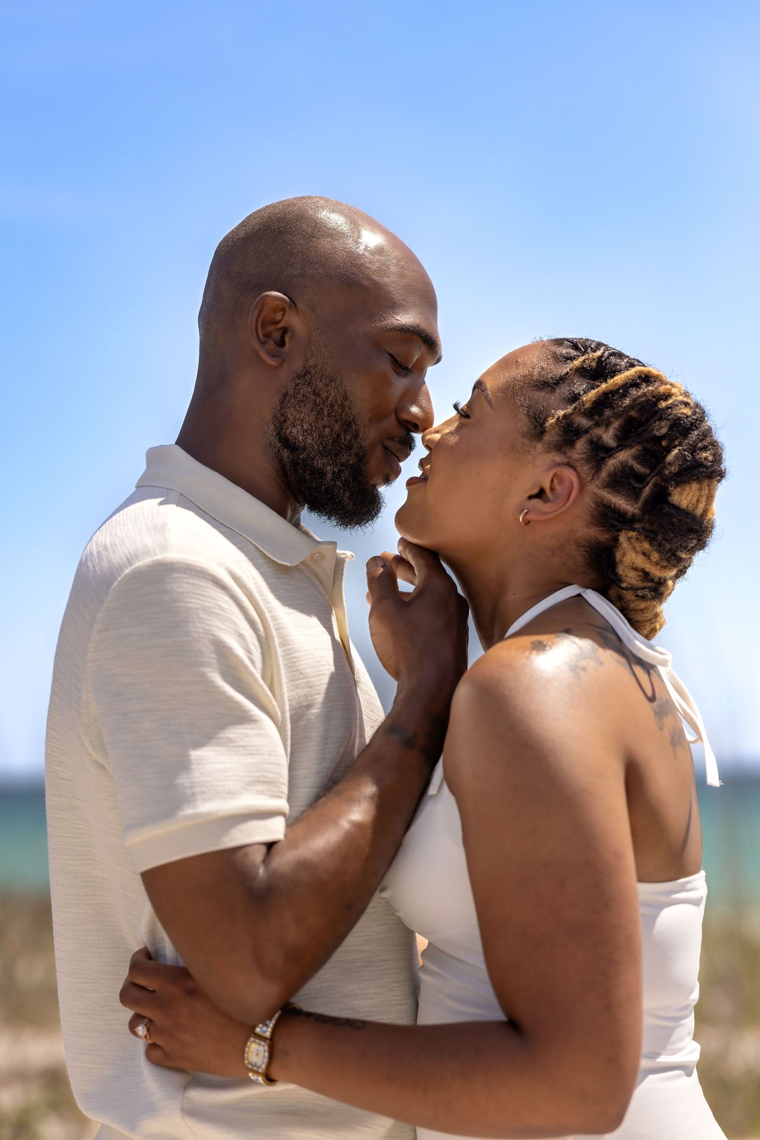 A romantic Black couple sharing an intimate moment on a sunny beach, dressed in white outfits.