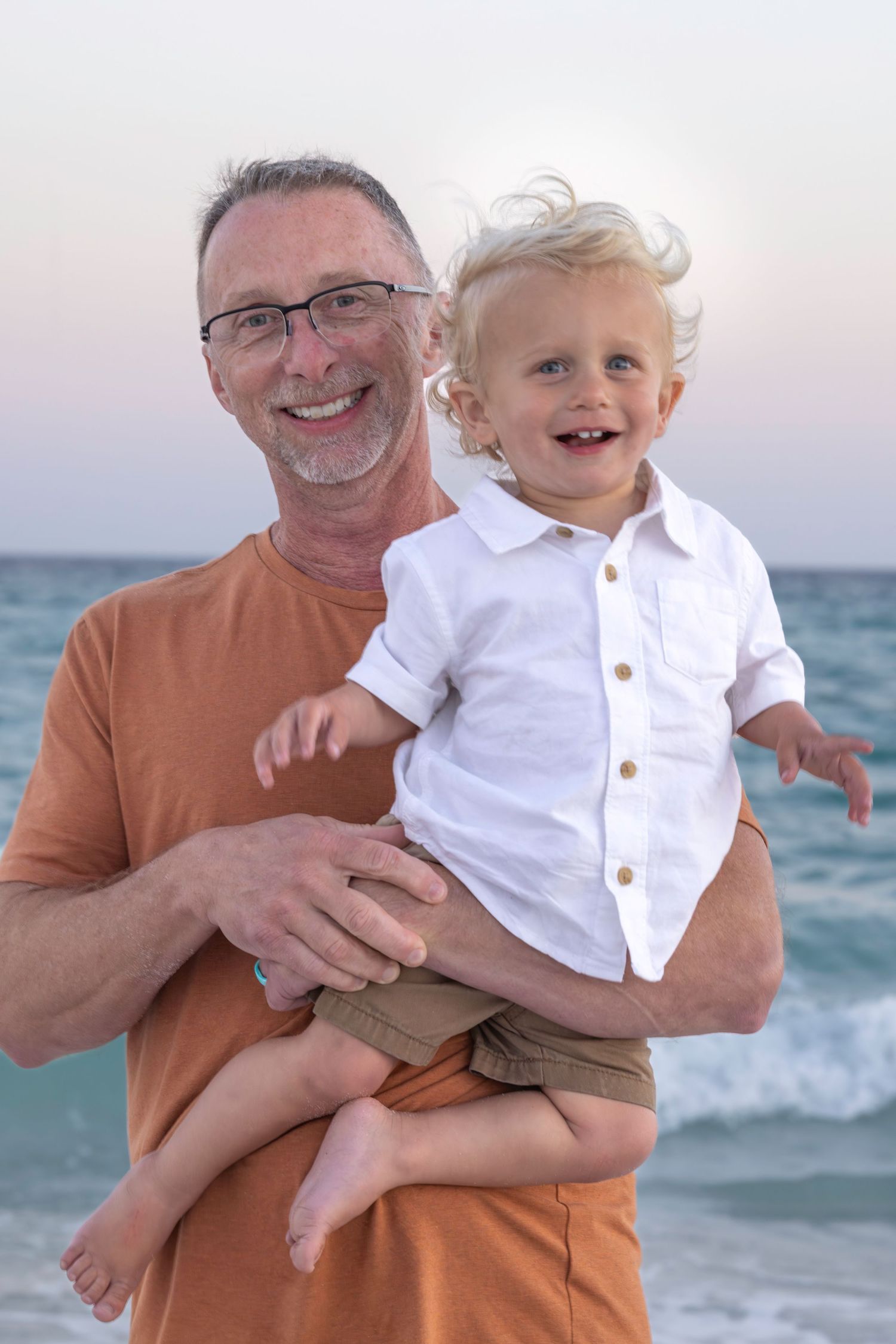 A cheerful grandfather wearing glasses holds a laughing blond toddler boy on a beach at dusk.