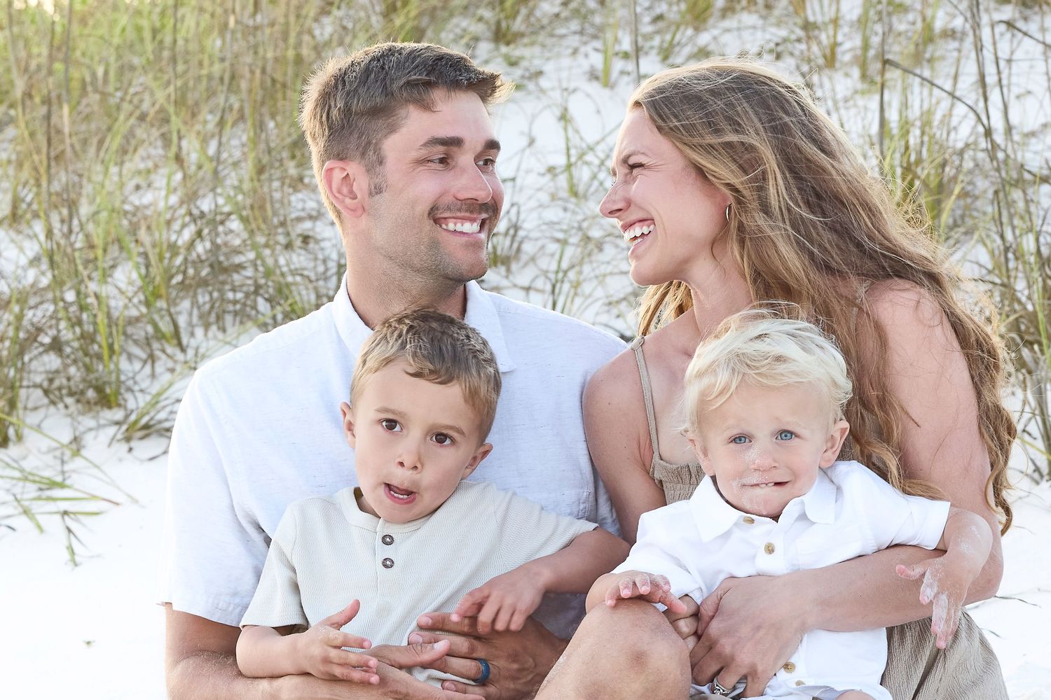 Happy family of four smiling together on a sandy beach, parents with two young blonde boys in white outfits.