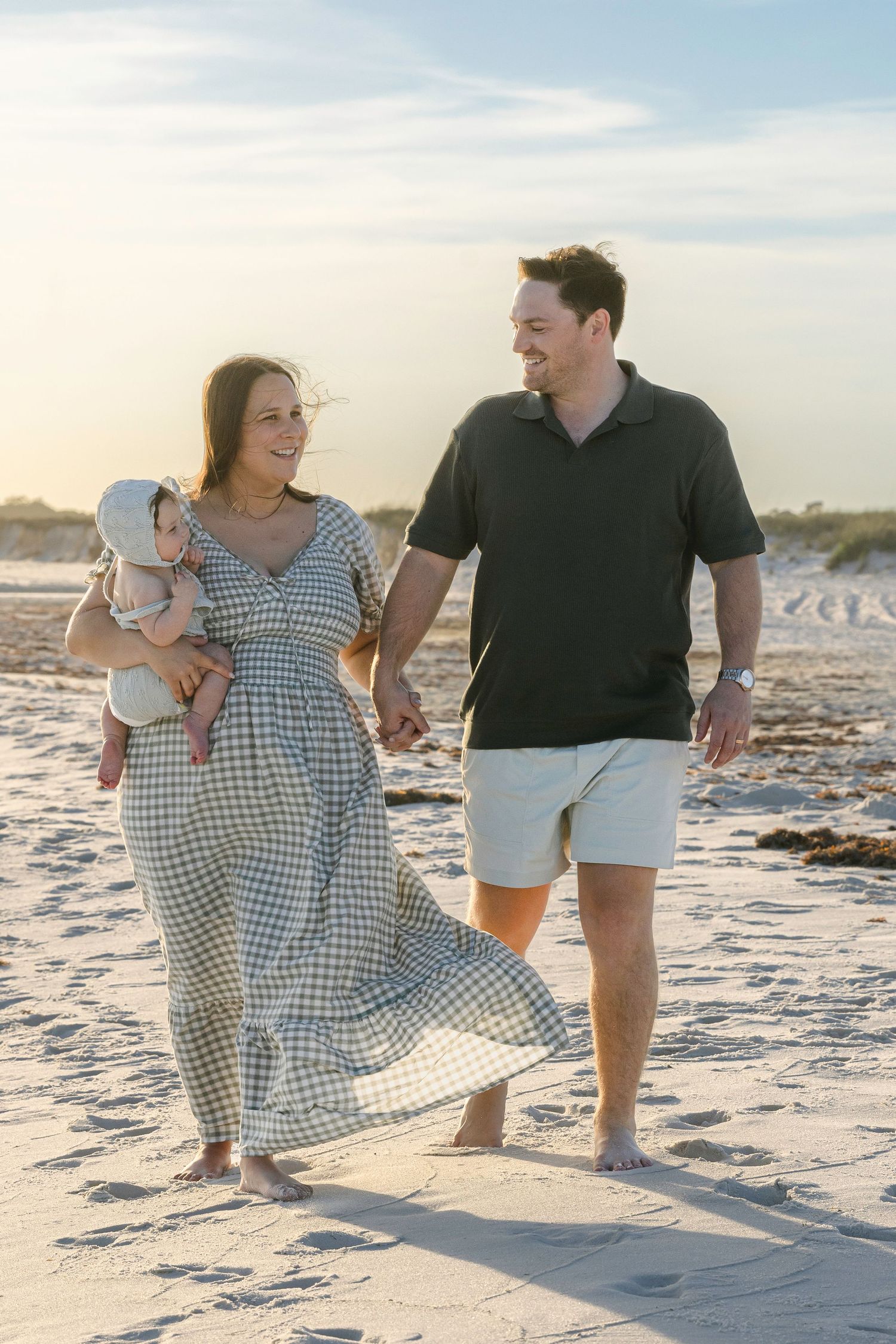 Happy young family with baby walking on sandy beach at sunset, holding hands in casual summer outfits.