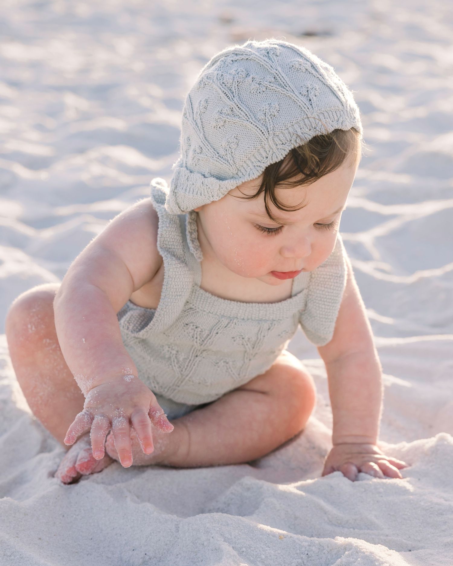A baby in a knit bonnet and romper sits on sandy beach, exploring the sand with chubby hands.